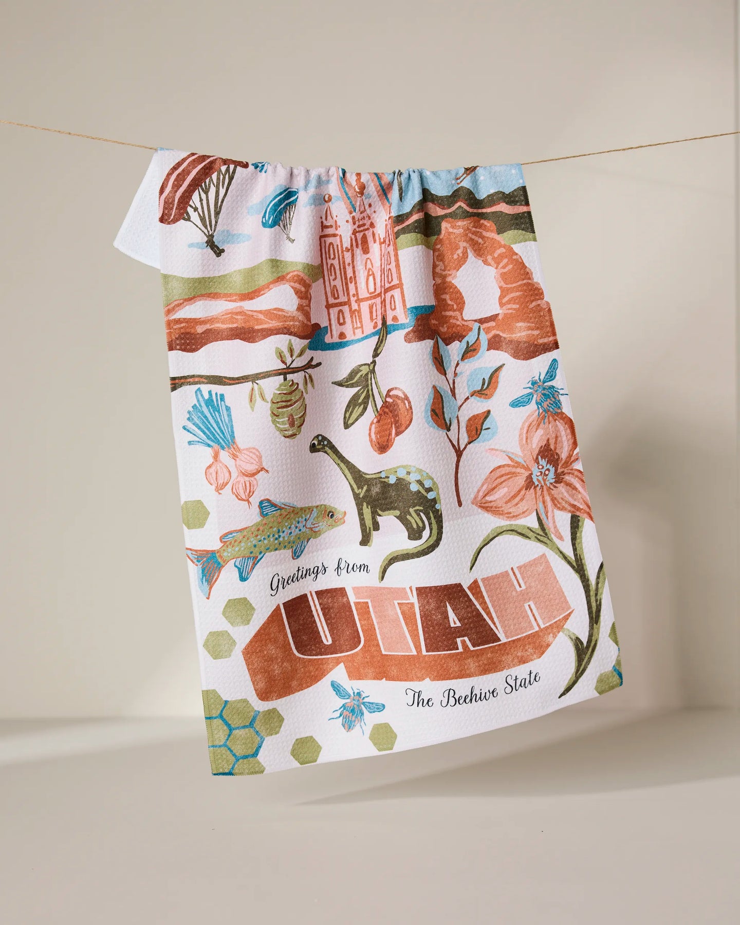 The Geometry Greetings From Utah tea towel features Utah-inspired illustrations—mountains, birds, cherries, and the Salt Lake Temple—with “Greetings from UTAH The Beehive State,” made from recycled materials.