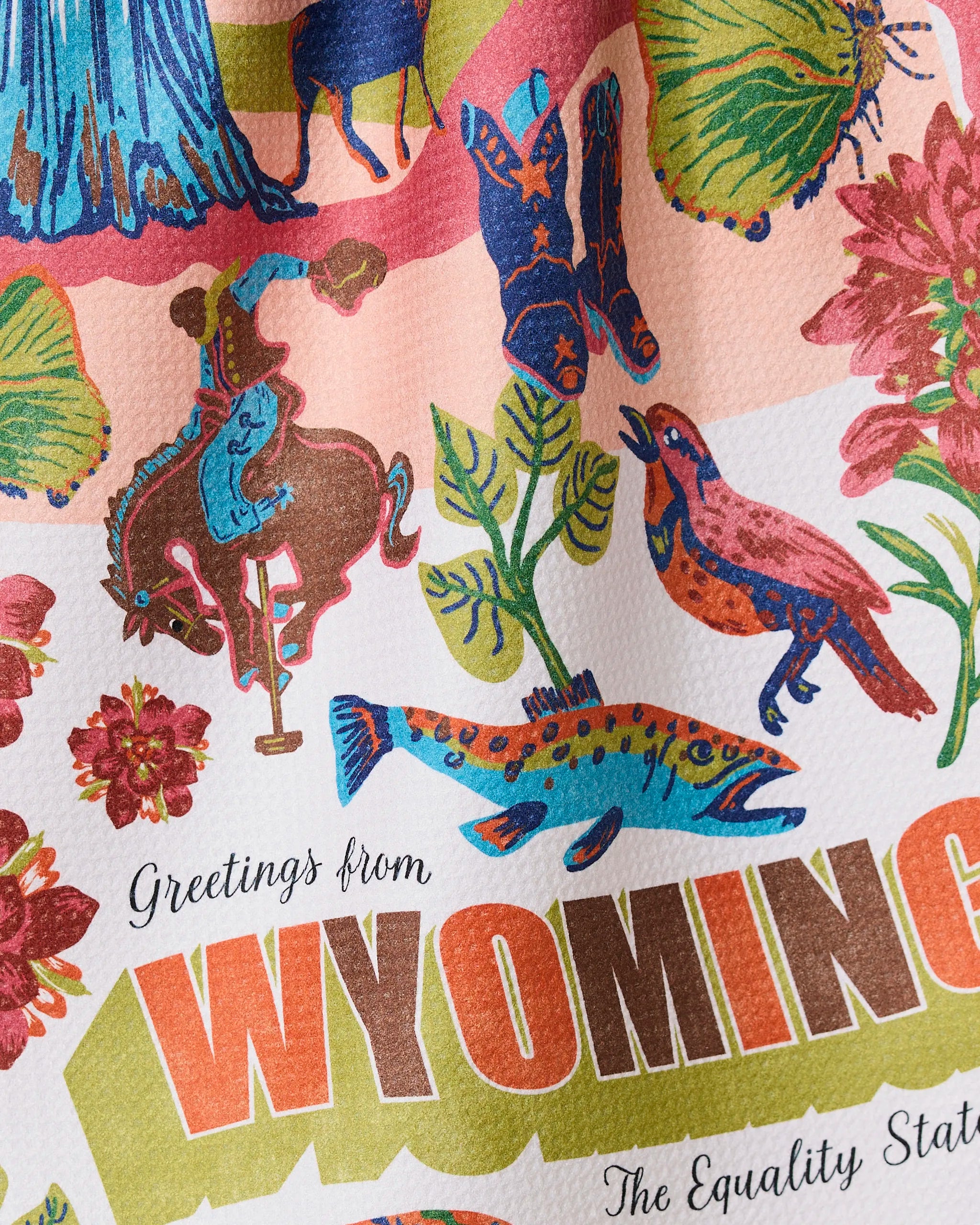 The Geometry Greetings From Wyoming ultra-absorbent towel showcases a colorful cowboy, horse, flowers, bird, fish, and the text Greetings from WYOMING with The Equality State on a textured background.