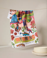The Geometry “Greetings From Wyoming” recycled kitchen towel, featuring vibrant illustrations of local wildlife, flowers, and landscapes plus the phrase “Greetings from WYOMING The Equality State,” hangs above stacked white plates.