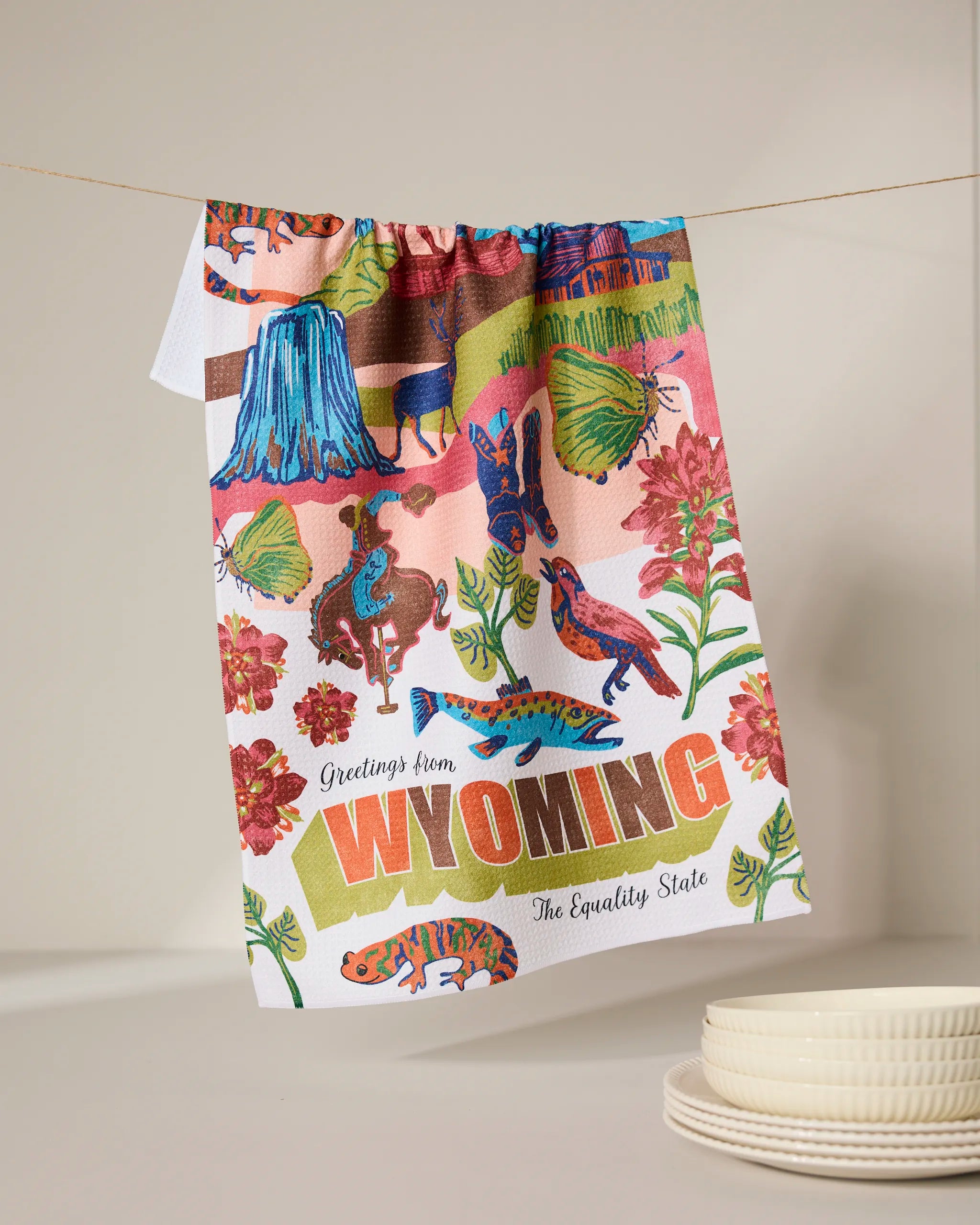 The Geometry “Greetings From Wyoming” recycled kitchen towel, featuring vibrant illustrations of local wildlife, flowers, and landscapes plus the phrase “Greetings from WYOMING The Equality State,” hangs above stacked white plates.
