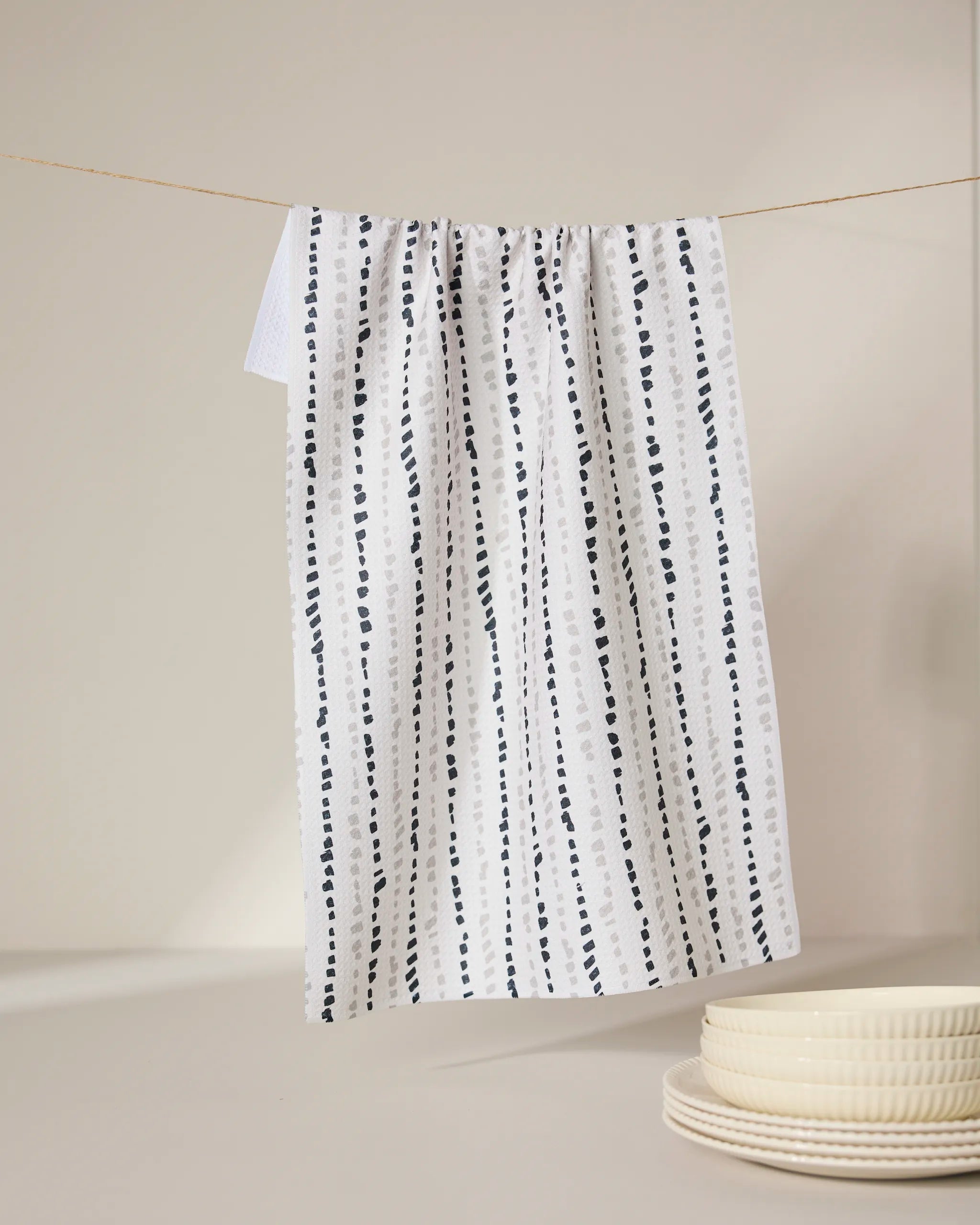 A GEOMETRY Grey Prints recycled kitchen towel with vertical dotted lines hangs on a string, with stacked cream-colored plates in the foreground on a neutral background.