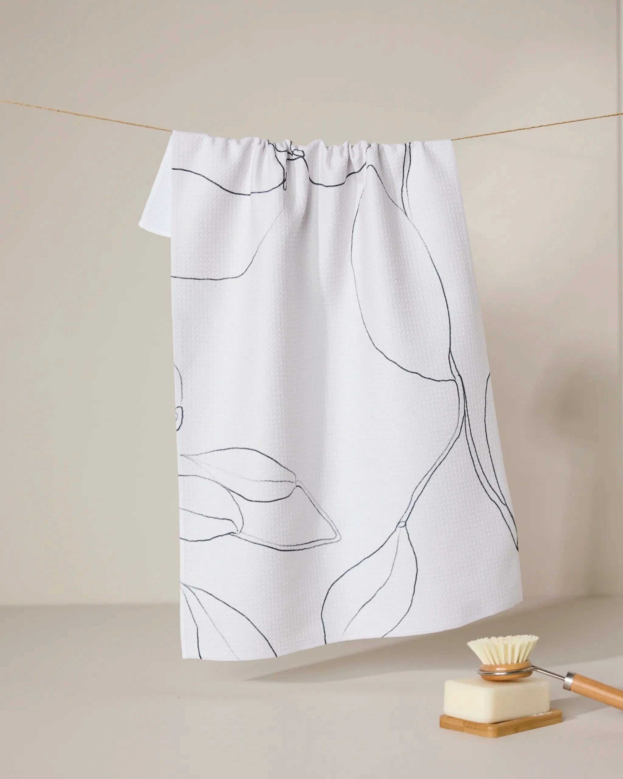 A GEOMETRY Grown recycled white kitchen towel with a black leaf pattern hangs on a string, while a natural bristle dish brush with a wooden handle rests nearby against a neutral, minimalistic background.