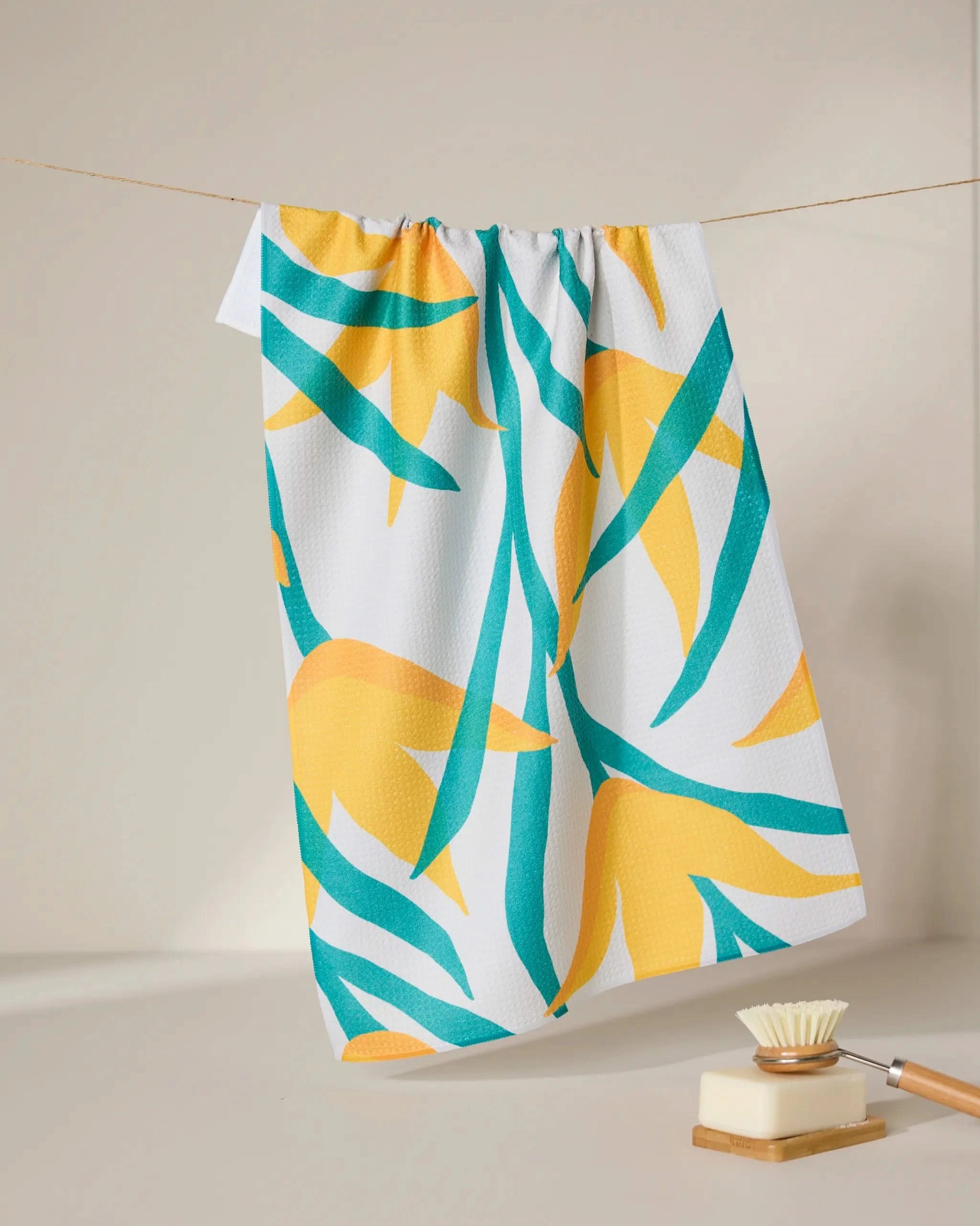 The GEOMETRY Growth tea towel, featuring a yellow and teal leaf pattern, hangs on a clothesline. Below, a wooden-handled dish brush rests on a beige surface, all set against a simple neutral background.