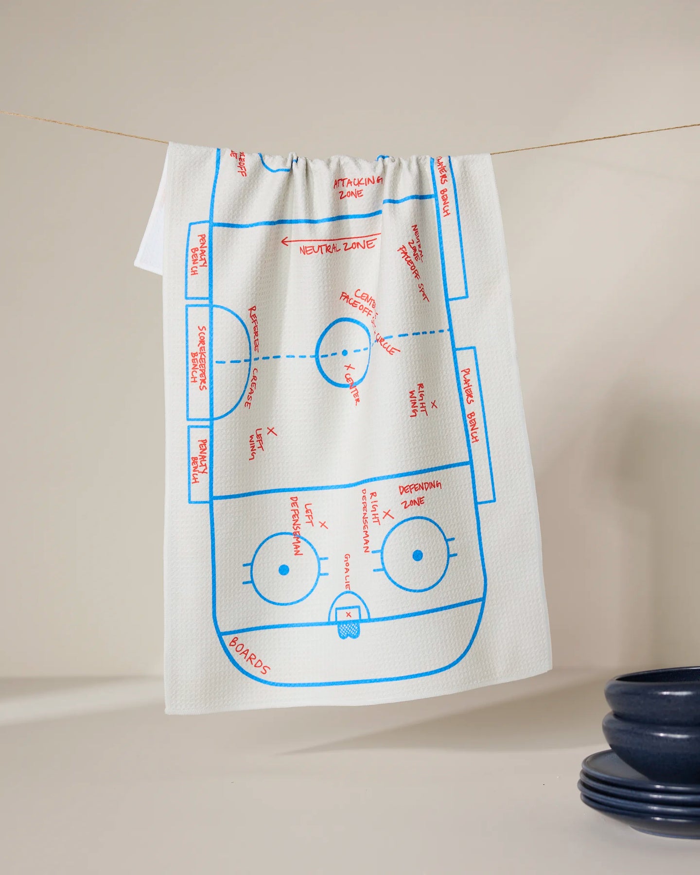 The GEOMETRY Hockey kitchen towel, in white, hangs on a line and displays a blue and red ice hockey rink diagram with labeled zones and markings; dark blue bowls sit in the lower right corner.
