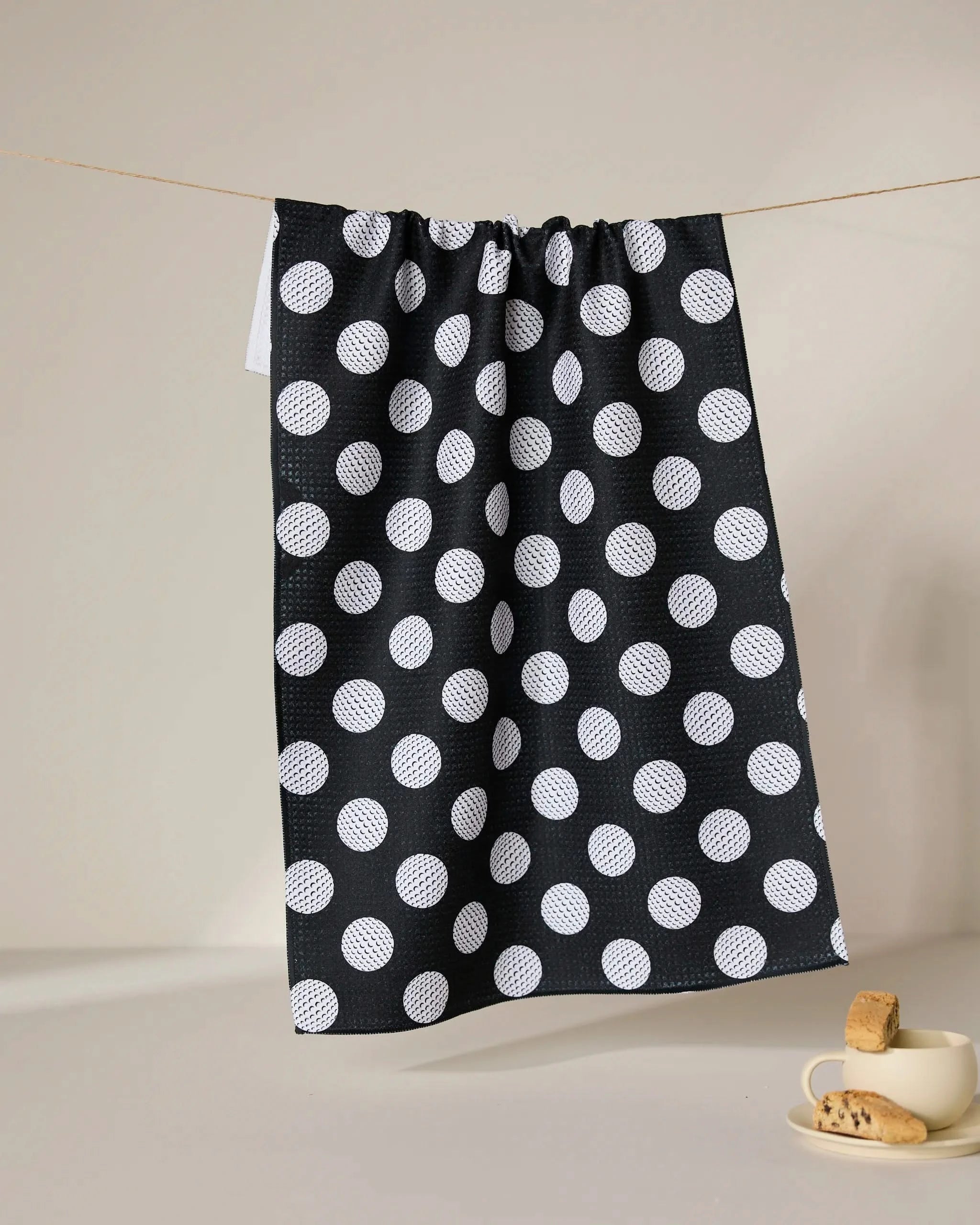 The GEOMETRY Hole In One ultra-absorbent tea towel, featuring large white polka dots on black, hangs on a clothesline; nearby, a white cup with saucer and cookies highlights the towel’s perfect mix of charm and practicality.
