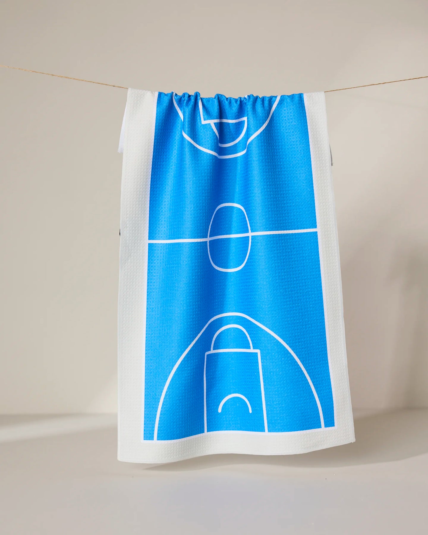 The Hoops towel by GEOMETRY, ultra-absorbent and white with a vibrant blue basketball court design, hangs on a clothesline against a plain, light background.