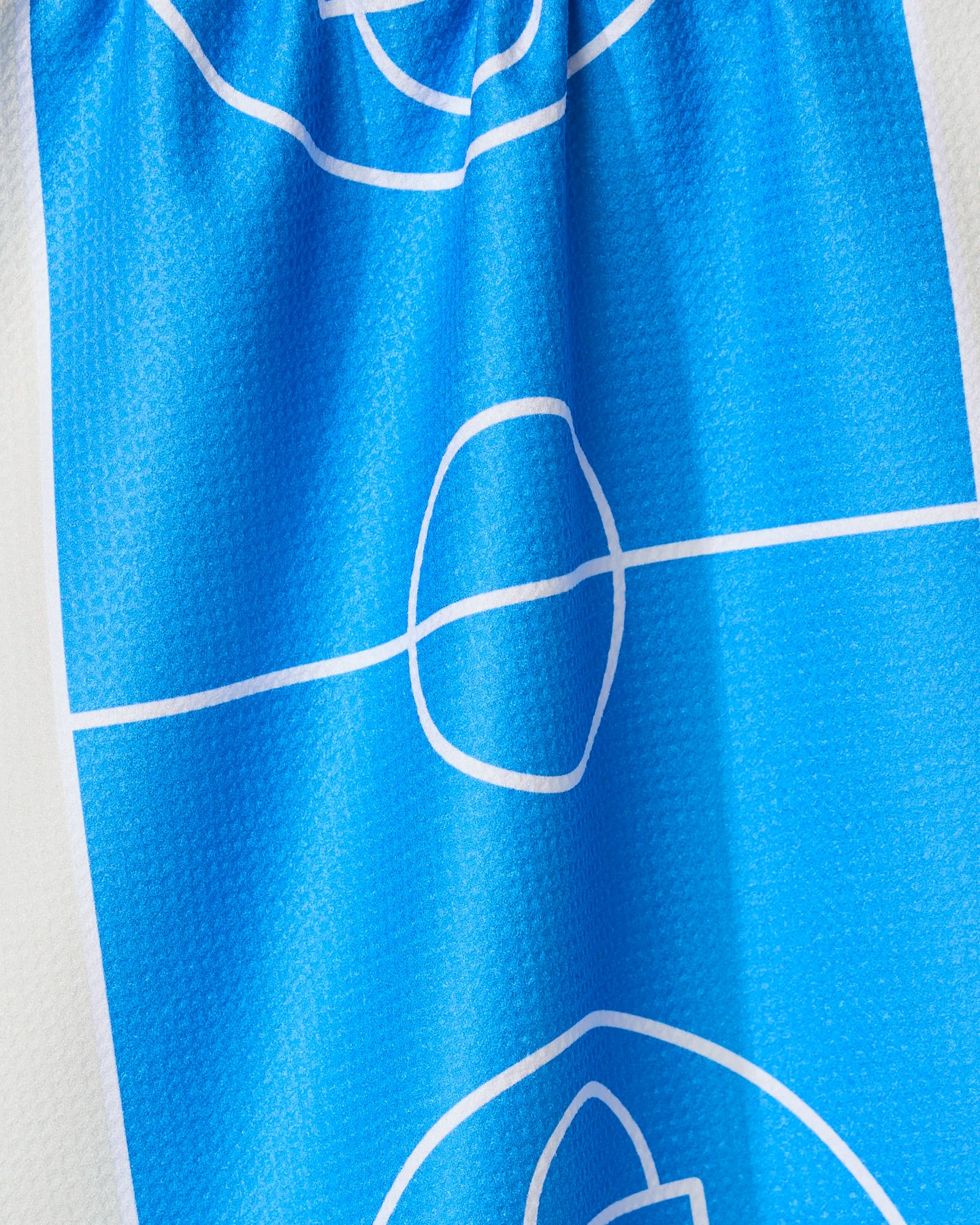 A close-up of the GEOMETRY Hoops tea towel shows its blue fabric with a white soccer field design; the ultra-absorbent towel is slightly wrinkled.
