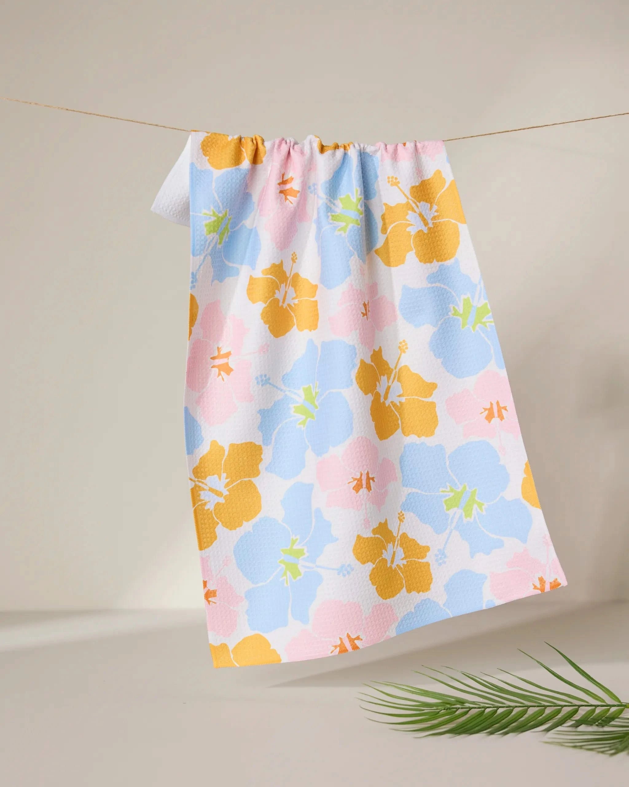 The GEOMETRY Hula Hues dish towel, featuring a pastel floral pattern in yellow, blue, and pink, hangs on a thin string against a neutral background with a green palm leaf in the foreground.