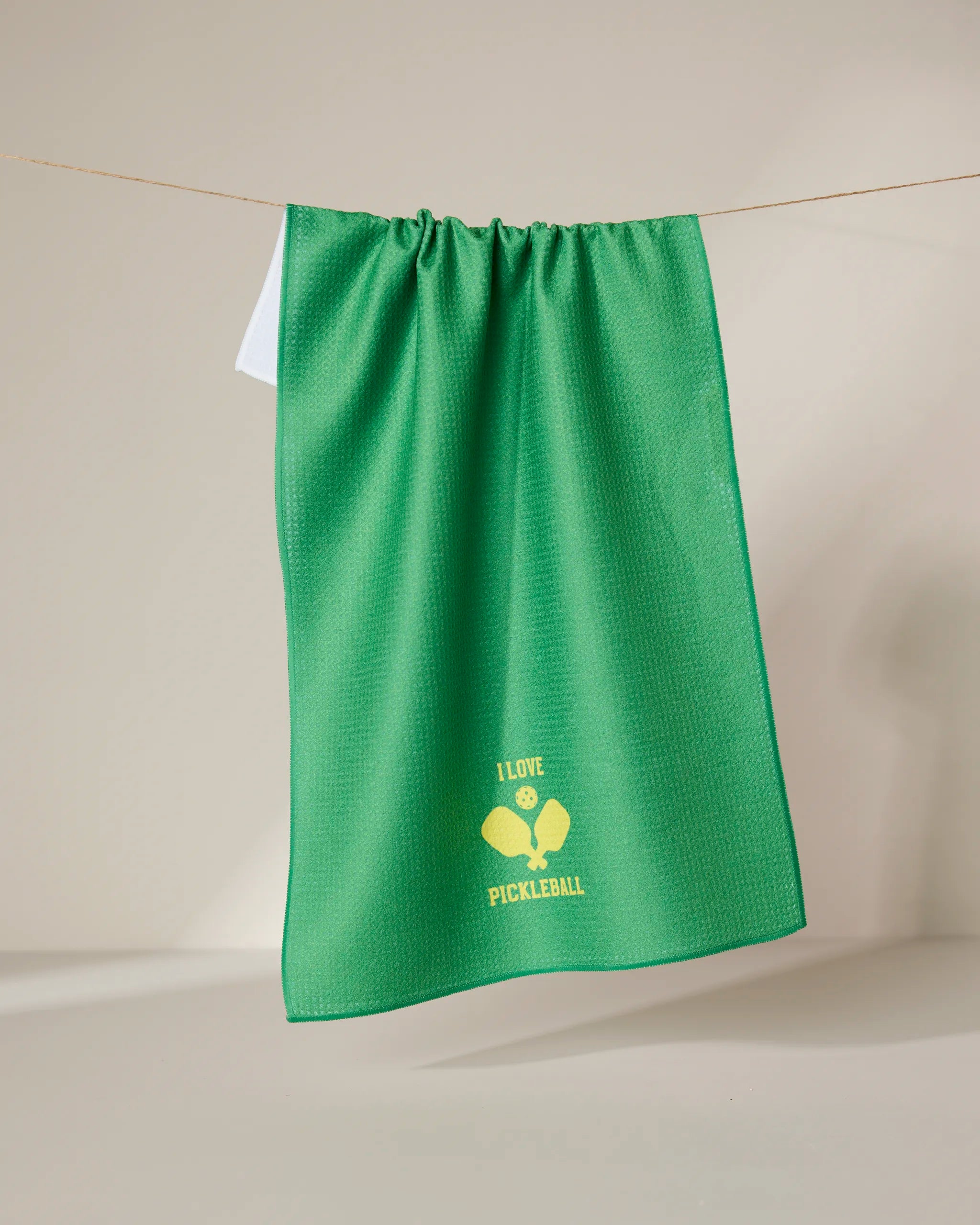 The GEOMETRY I Love Pickleball green tea towel, made from recycled materials, hangs on a line with a yellow graphic of two crossed pickleball paddles, a ball, and “I LOVE PICKLEBALL” near the bottom center.