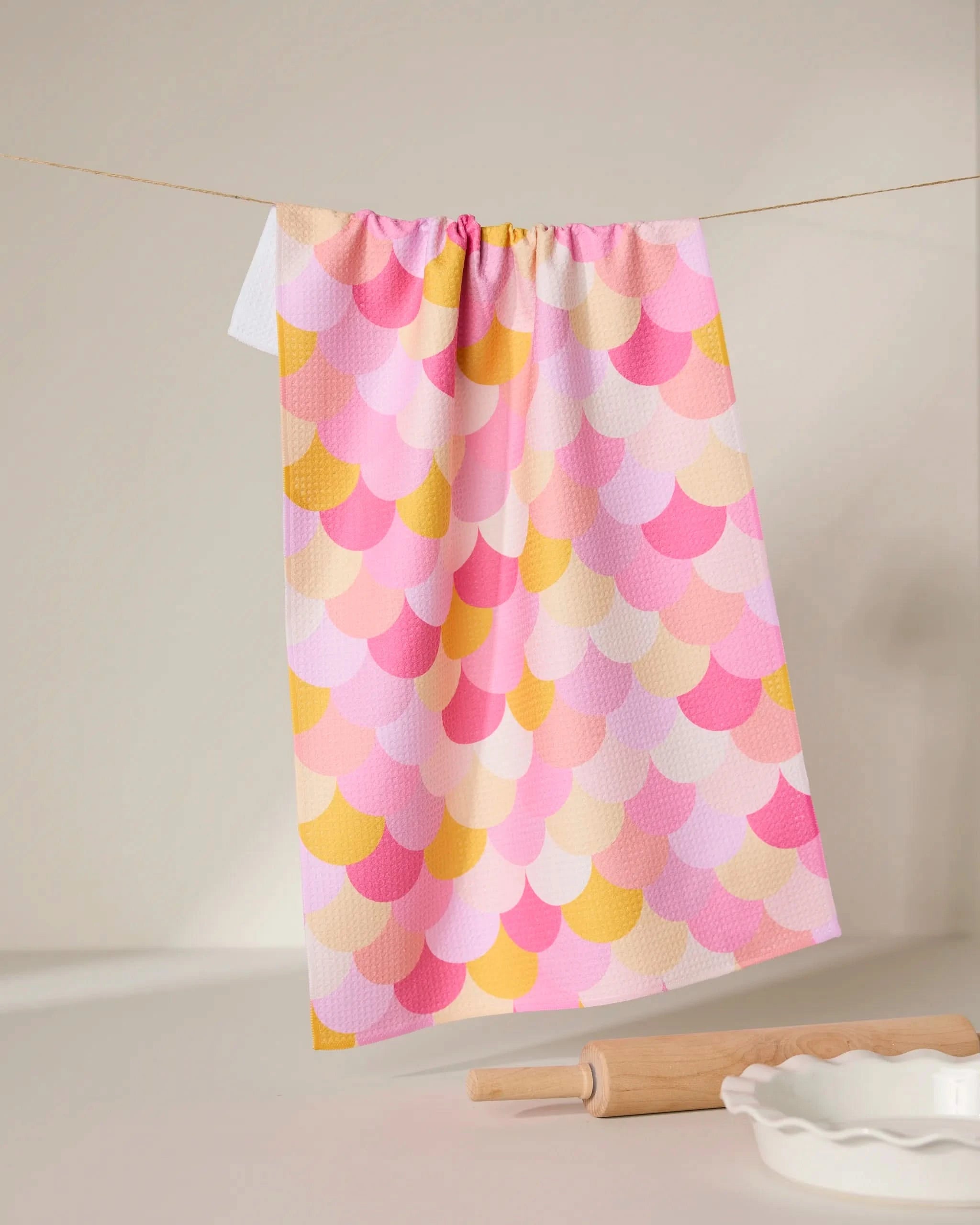 The GEOMETRY Inherit dish towel, featuring a pastel scallop pattern in pink, yellow, and orange, hangs on a string. A rolling pin and white ceramic dish sit on the white surface in the foreground.