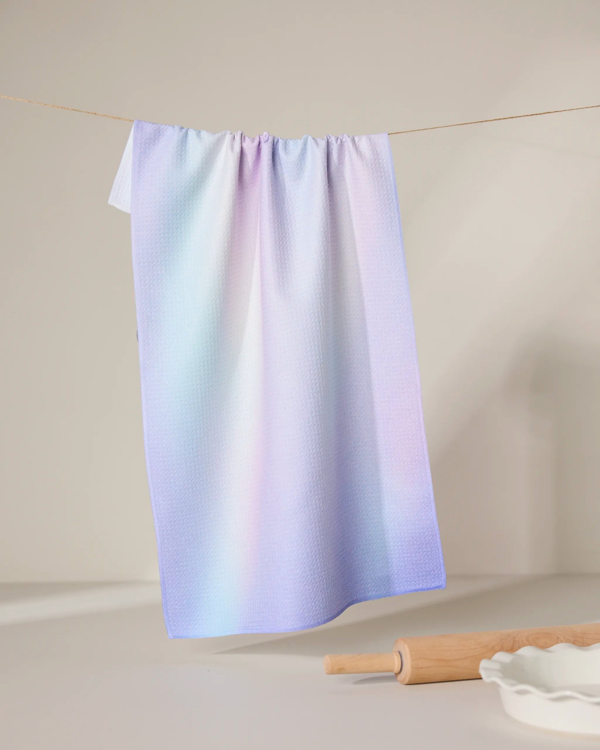 The GEOMETRY Iridescence kitchen towel, featuring an ultra-absorbent pastel gradient in pink, blue, and purple, hangs on a line. Below are a wooden rolling pin and a white fluted dish on a light surface.