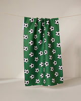 The GEOMETRY Kick Off eco-friendly kitchen towel in green features a repeating black and white soccer ball pattern and hangs on a line against a plain, light-colored background.