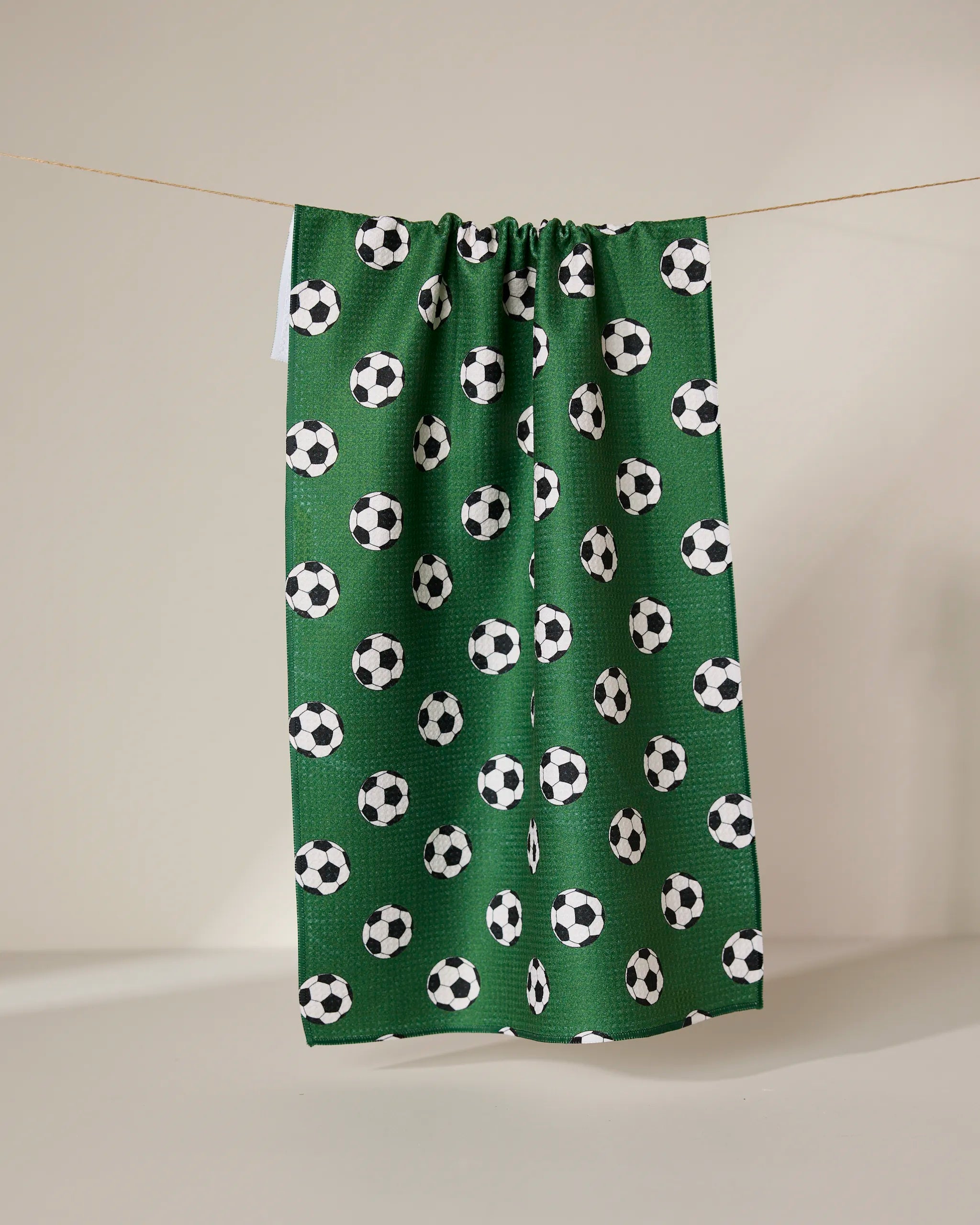 The GEOMETRY Kick Off eco-friendly kitchen towel in green features a repeating black and white soccer ball pattern and hangs on a line against a plain, light-colored background.
