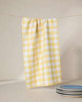 The GEOMETRY Lemon Gingham recycled kitchen towel hangs on a line with three blue and white patterned ceramic plates stacked nearby, all set against a neutral background.