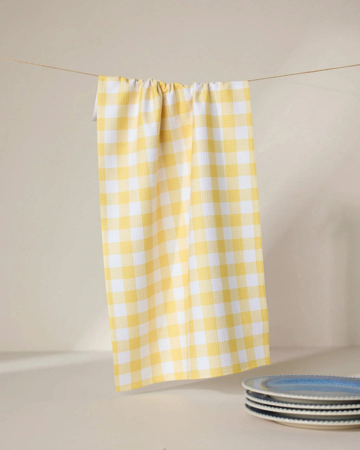 The GEOMETRY Lemon Gingham recycled kitchen towel hangs on a line with three blue and white patterned ceramic plates stacked nearby, all set against a neutral background.