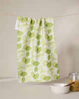 The GEOMETRY Limes tea towel with a green lemon and lime pattern hangs on a clothesline. Below, a white bowl with a wooden spoon and a glass jar of cereal or granola sit on a clean white surface.