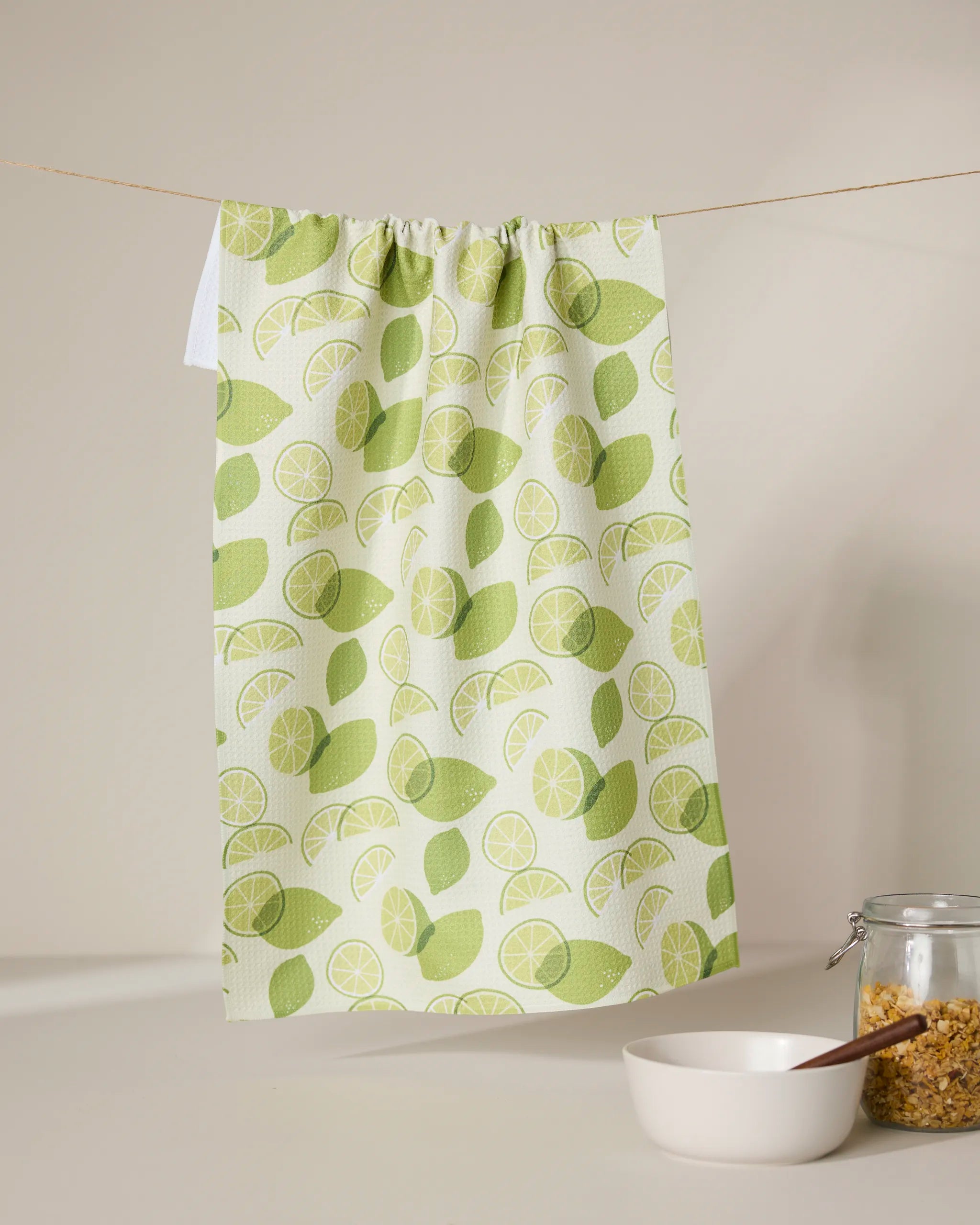 The GEOMETRY Limes tea towel with a green lemon and lime pattern hangs on a clothesline. Below, a white bowl with a wooden spoon and a glass jar of cereal or granola sit on a clean white surface.