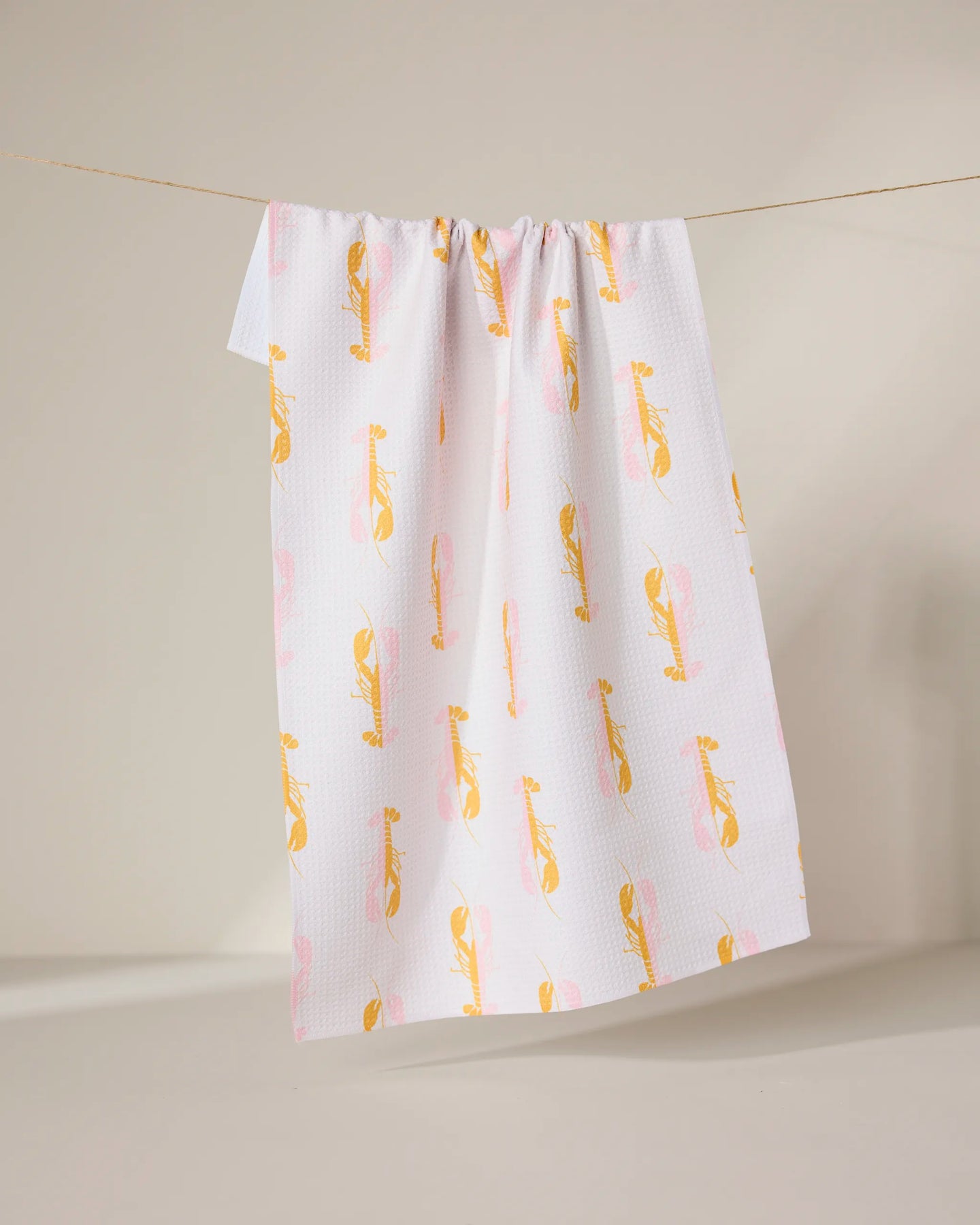 The GEOMETRY Lobster Love towel, made from recycled materials, features a white background with a yellow and pink abstract bird pattern and hangs on a clothesline against a neutral backdrop.
