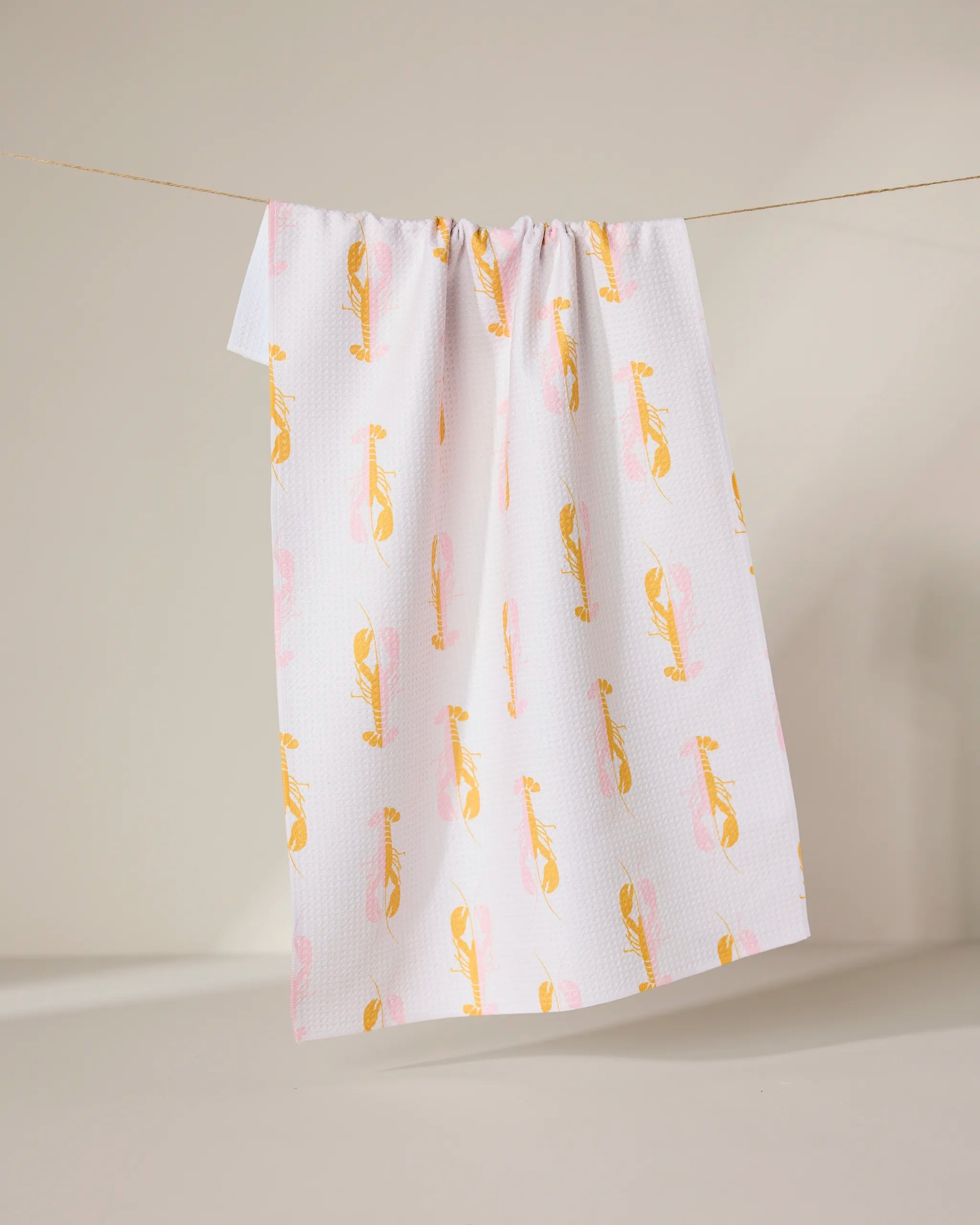 The GEOMETRY Lobster Love towel, made from recycled materials, features a white background with a yellow and pink abstract bird pattern and hangs on a clothesline against a neutral backdrop.