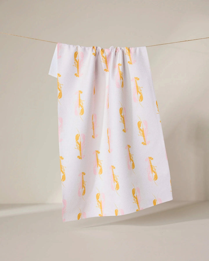 The GEOMETRY Lobster Love towel, made from recycled materials, features a white background with a yellow and pink abstract bird pattern and hangs on a clothesline against a neutral backdrop.