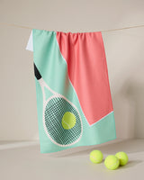 The GEOMETRY Love 15 tea towel, featuring a tennis racket and ball design in pastel green, coral, and white Geoweave fabric, hangs on a line with three yellow tennis balls beneath it.
