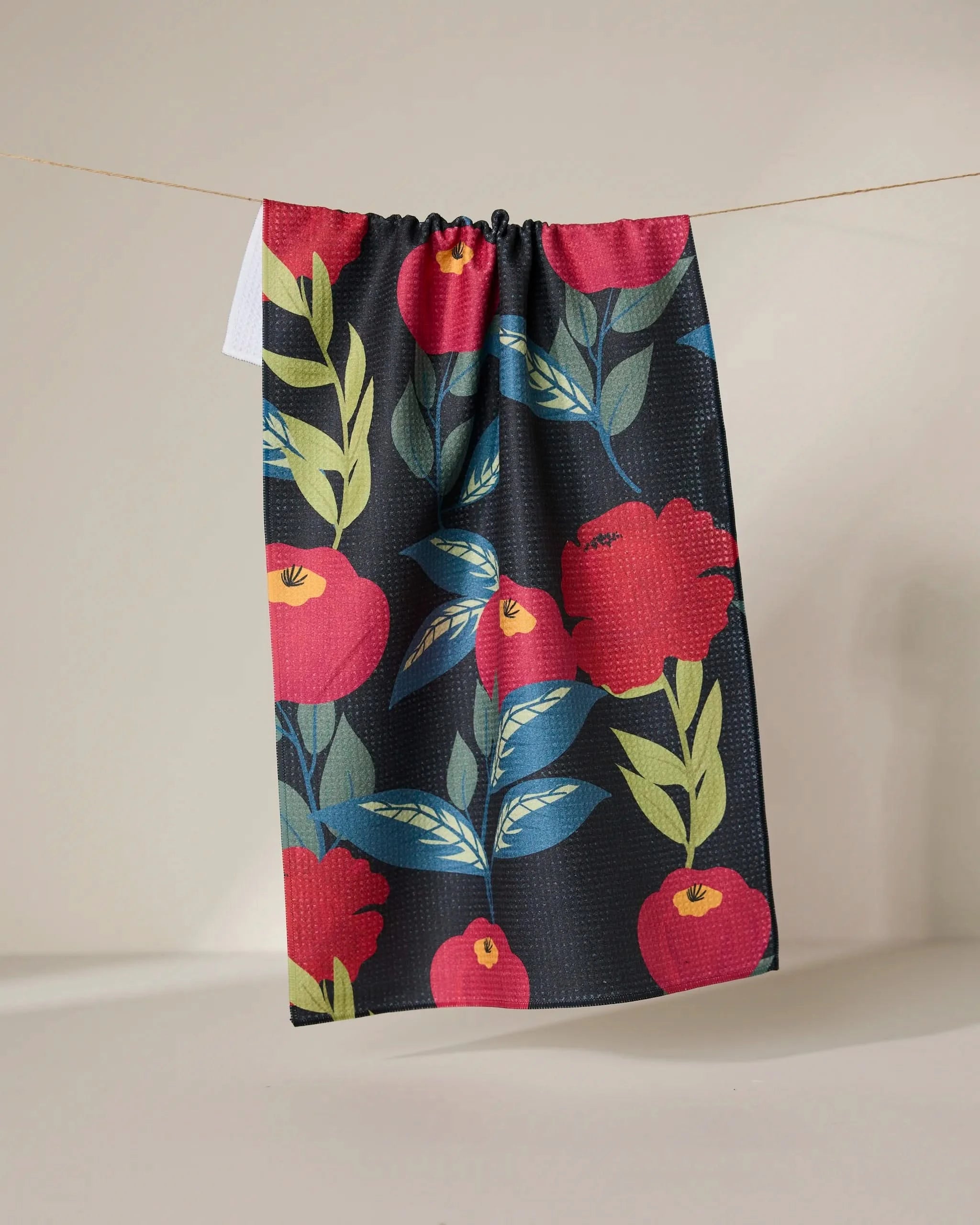 The GEOMETRY Lovely Night tea towel, featuring a bold red and yellow floral and leaf pattern on black, hangs on a string against a neutral background.