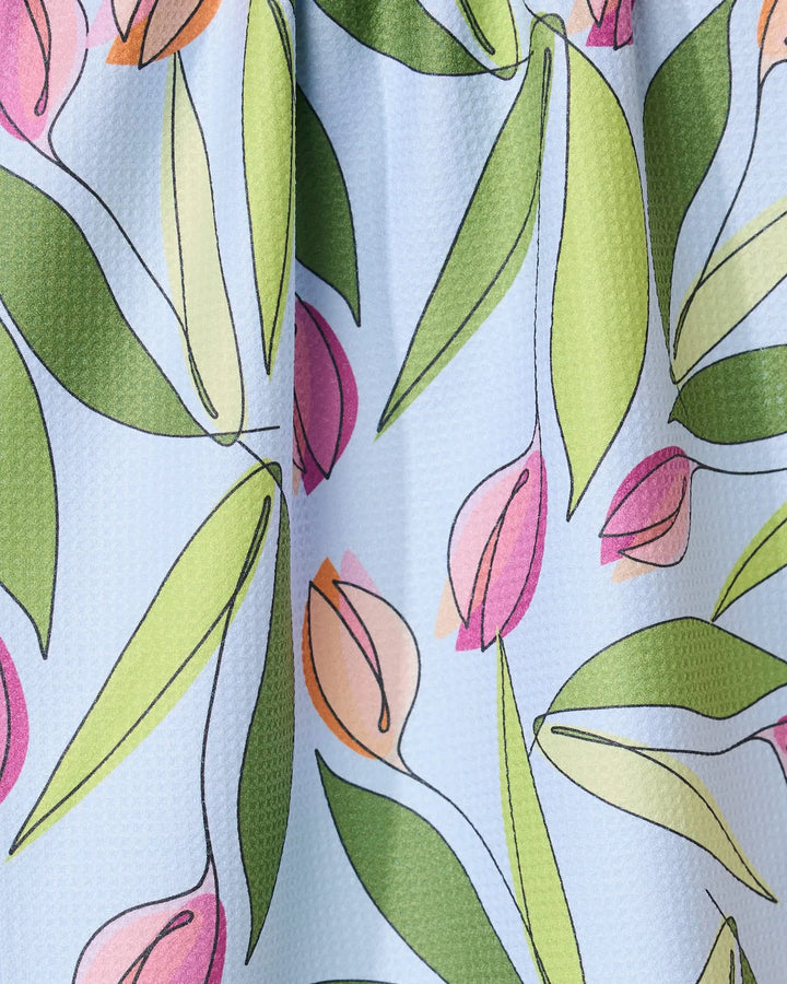A close-up of the GEOMETRY May Flowers absorbent quick-dry towel, showcasing pink and peach tulip-style flowers with green leaves on a textured light blue background.
