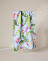 The GEOMETRY May Flowers recycled kitchen towel, featuring a pink and green tulip and leaf pattern on light blue, hangs from a string against a neutral backdrop.