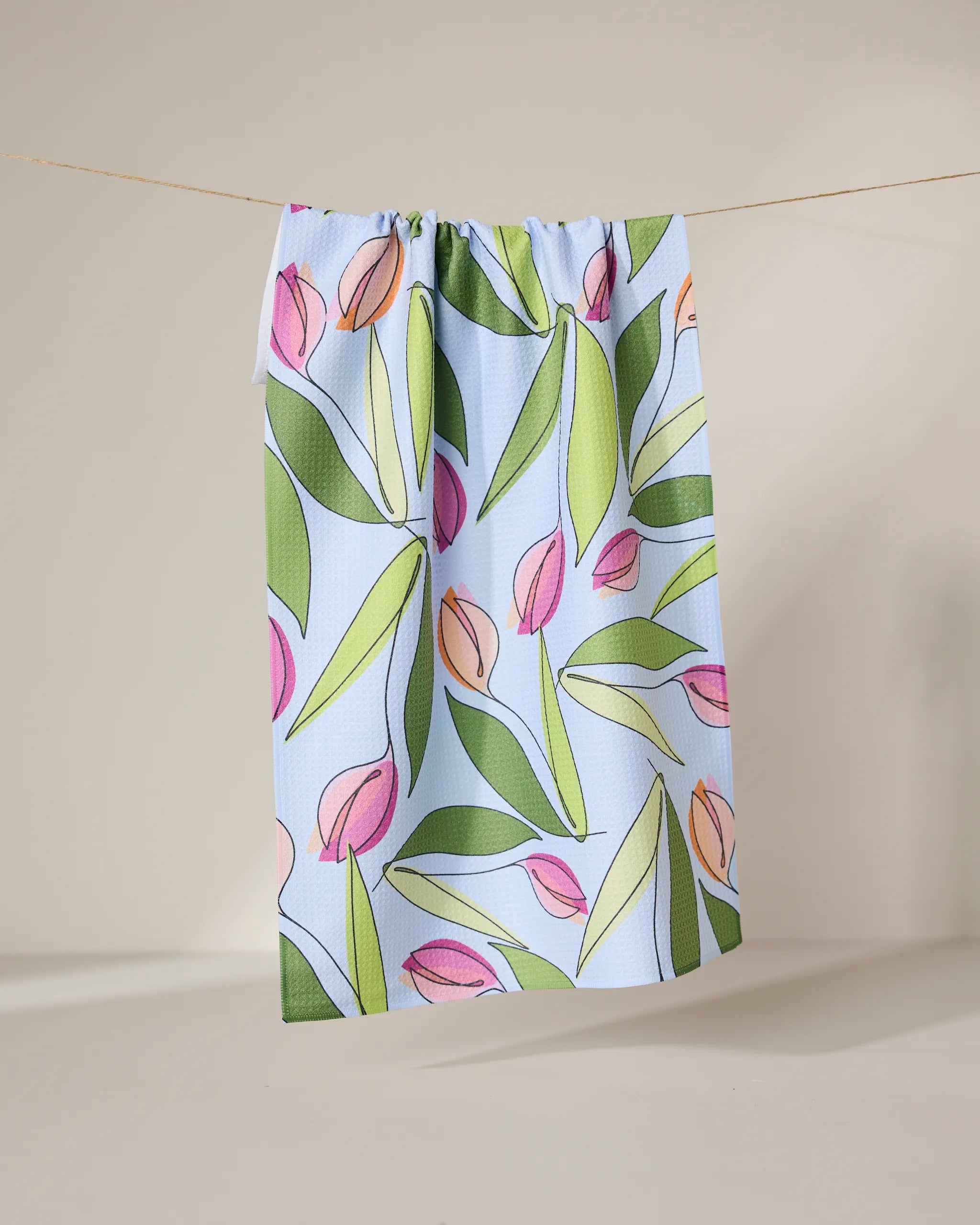 The GEOMETRY May Flowers recycled kitchen towel, featuring a pink and green tulip and leaf pattern on light blue, hangs from a string against a neutral backdrop.