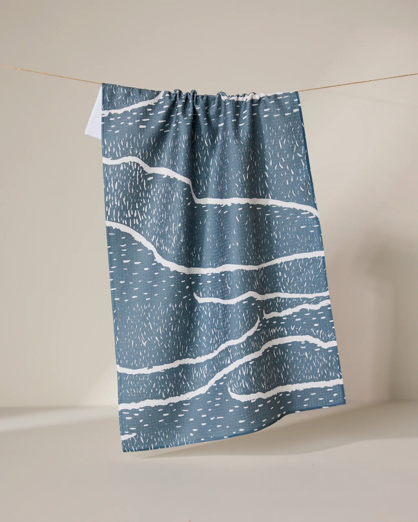 The GEOMETRY Wetlands - Midnight tea towel, featuring abstract wavy white lines and dashes on a blue base, hangs from a string against a neutral background. This absorbent dish towel combines style and functionality.