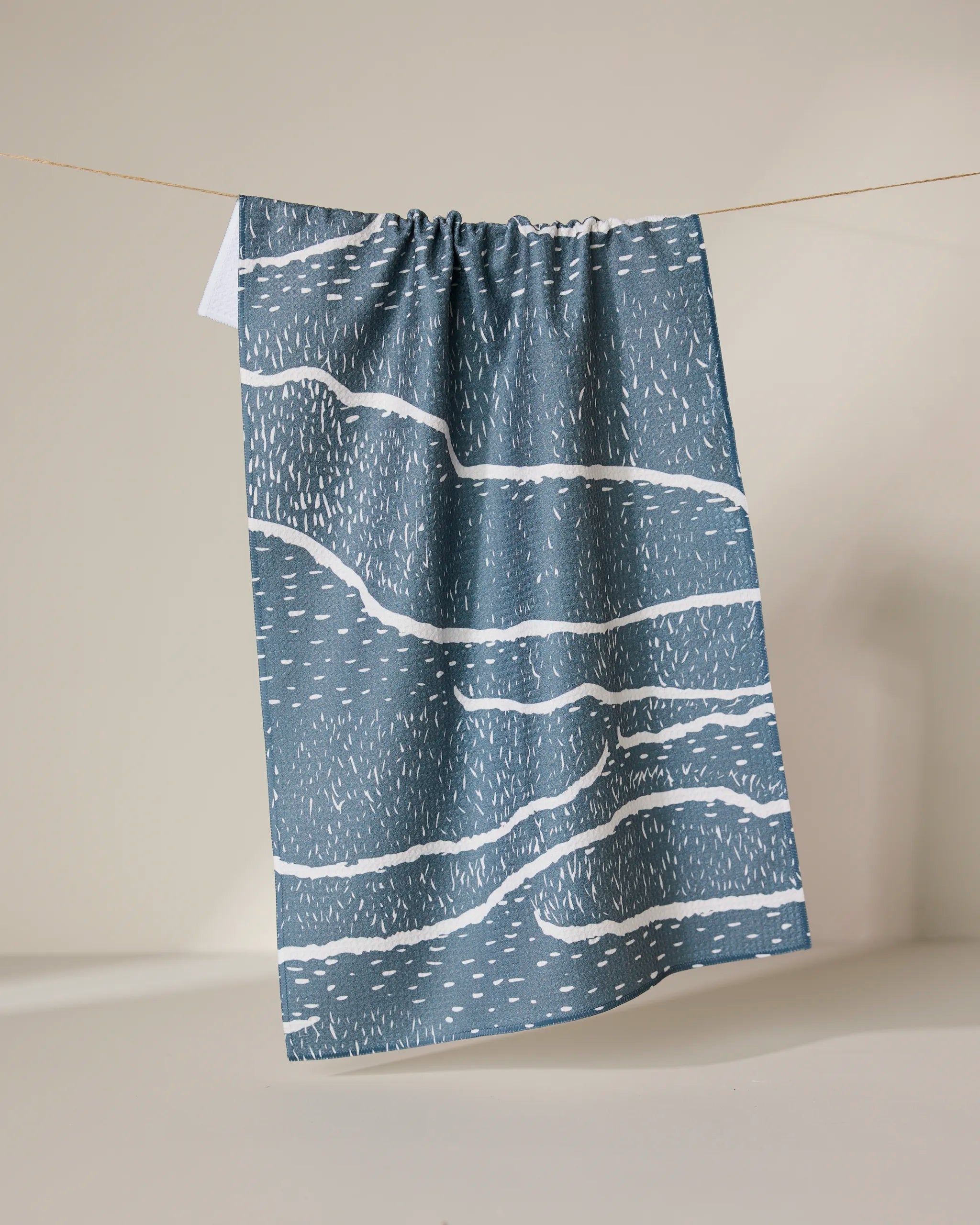 The GEOMETRY Wetlands - Midnight tea towel, featuring abstract wavy white lines and dashes on a blue base, hangs from a string against a neutral background. This absorbent dish towel combines style and functionality.