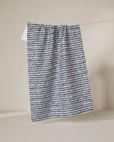 The Geometry Dot Dot Dot Fresh Linen recycled kitchen towel, featuring a black irregular dot pattern on white, hangs from a thin clothesline against a plain beige background.