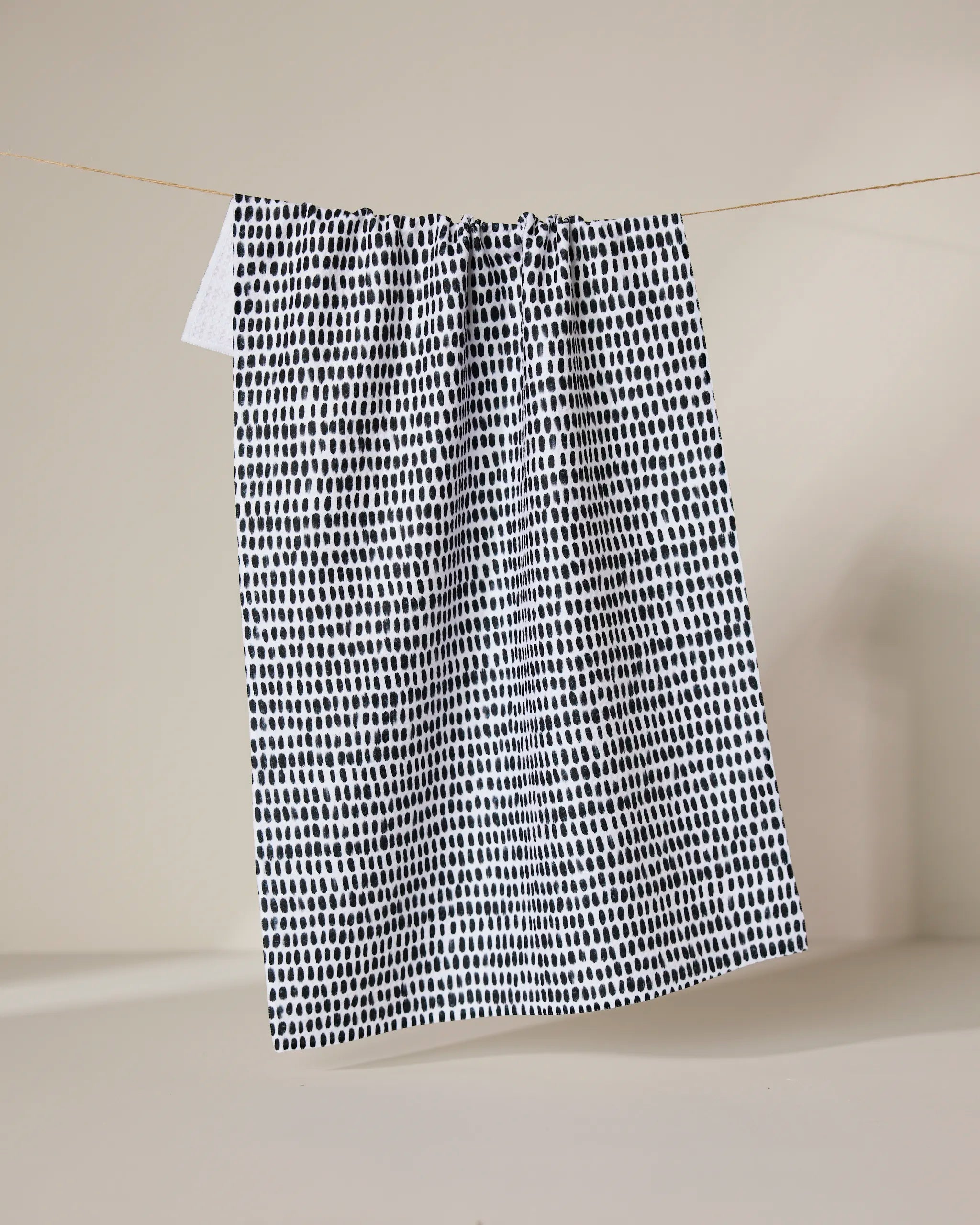 The Geometry Dot Dot Dot Fresh Linen recycled kitchen towel, featuring a black irregular dot pattern on white, hangs from a thin clothesline against a plain beige background.