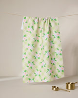 The GEOMETRY Doting and Devoted kitchen tea towel features a green and white checkered floral pattern, made from recycled materials. It hangs on a line above two gold measuring cups, with a neutral background that highlights its minimal charm.