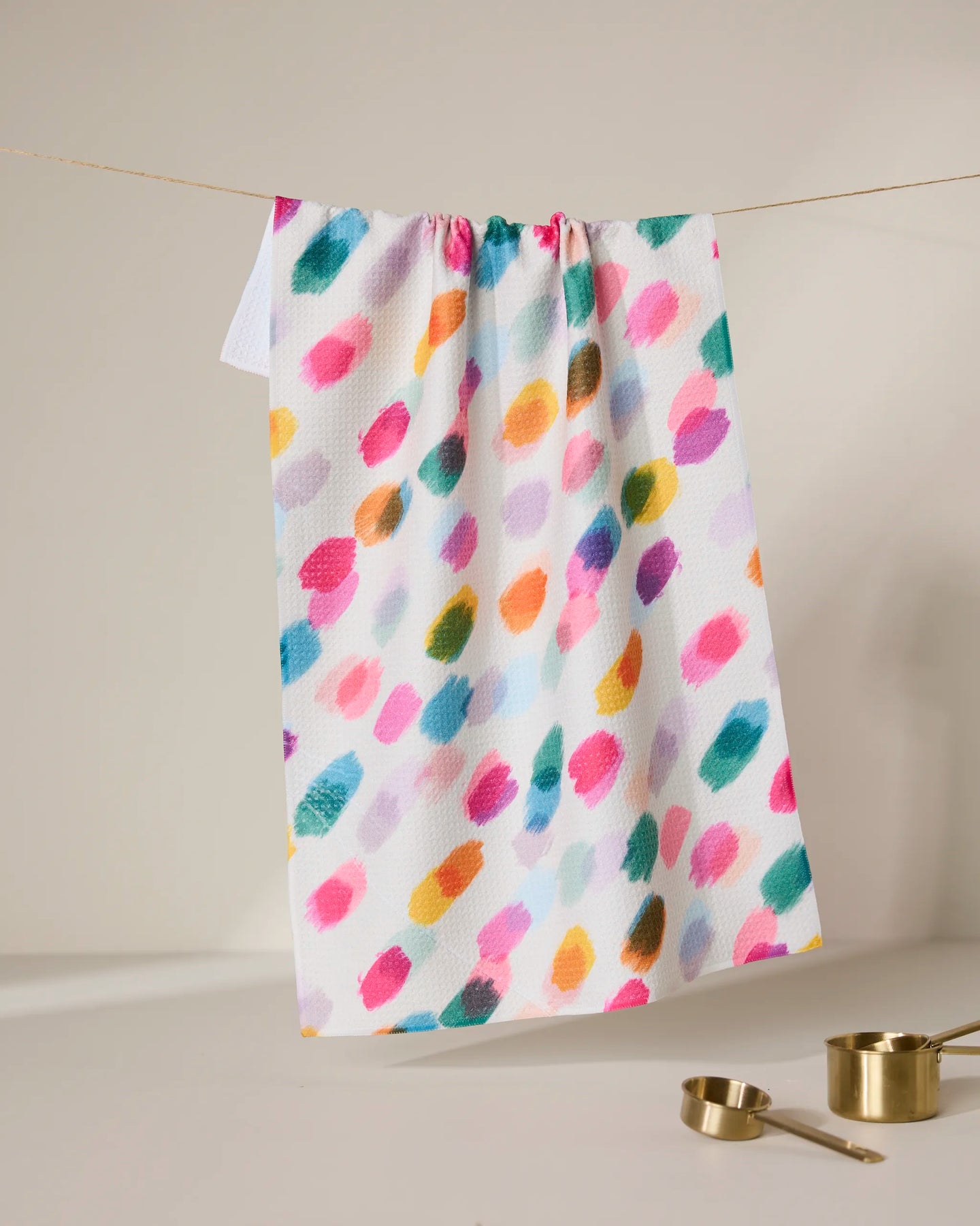 The GEOMETRY Downpour tea towel, made from recycled materials and featuring colorful, irregular brushstrokes, hangs on a softly lit line. Below, gold measuring cups and spoons rest on a neutral surface. Ultra-absorbent design.