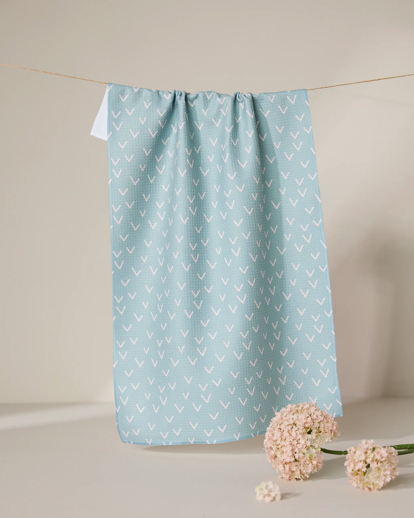 The GEOMETRY Drawn Doves tea towel, featuring a quick-drying light blue fabric with a white arrow pattern, hangs on a clothesline. Below it, light pink flowers are arranged on a beige surface against a neutral backdrop.