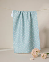 The GEOMETRY Drawn Doves tea towel, featuring a quick-drying light blue fabric with a white arrow pattern, hangs on a clothesline. Below it, light pink flowers are arranged on a beige surface against a neutral backdrop.