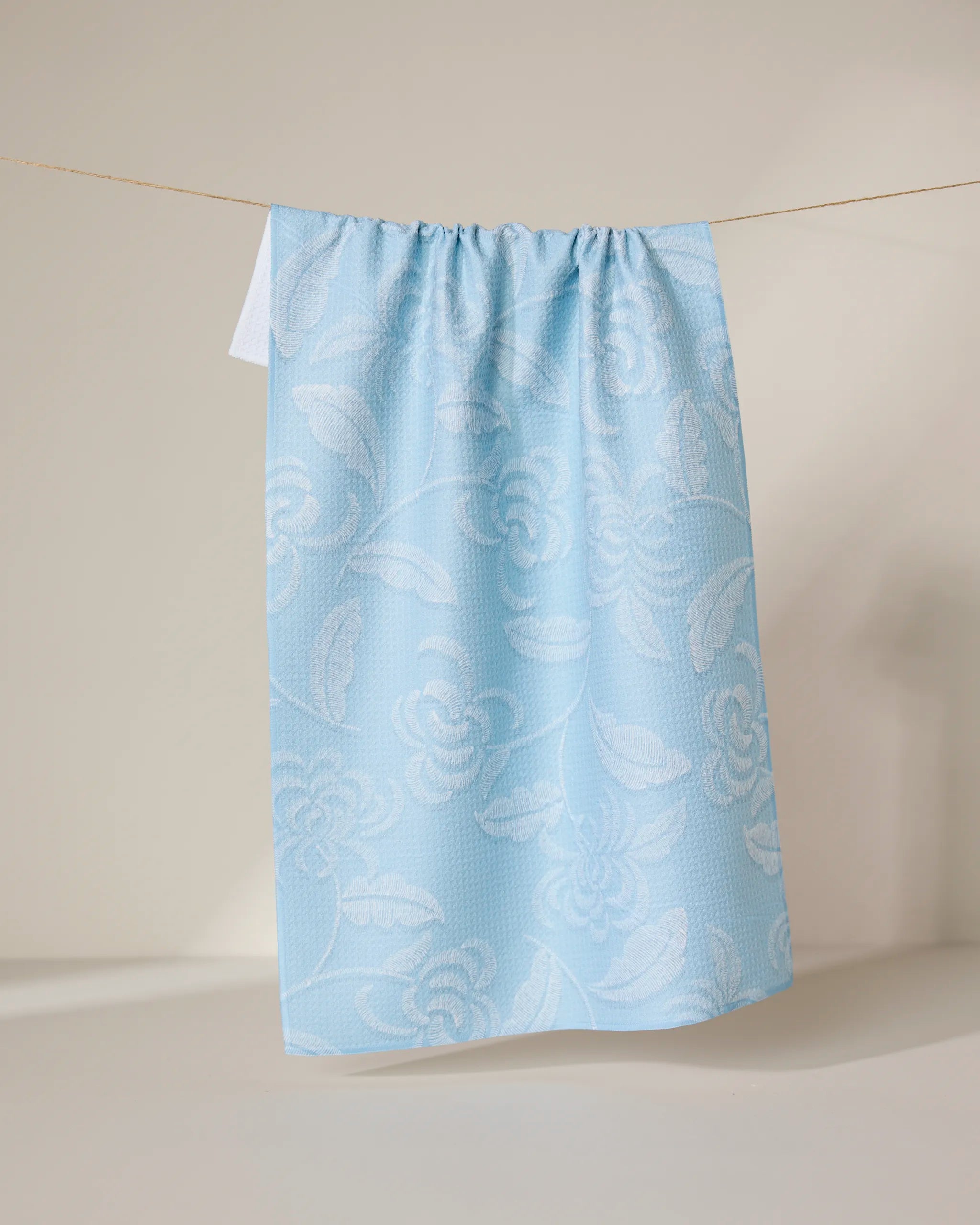 GEOMETRY’s Ediths Embroidery curtain features a light blue hue with white floral patterns, made from recycled materials, and is shown hanging by a string against a neutral background.