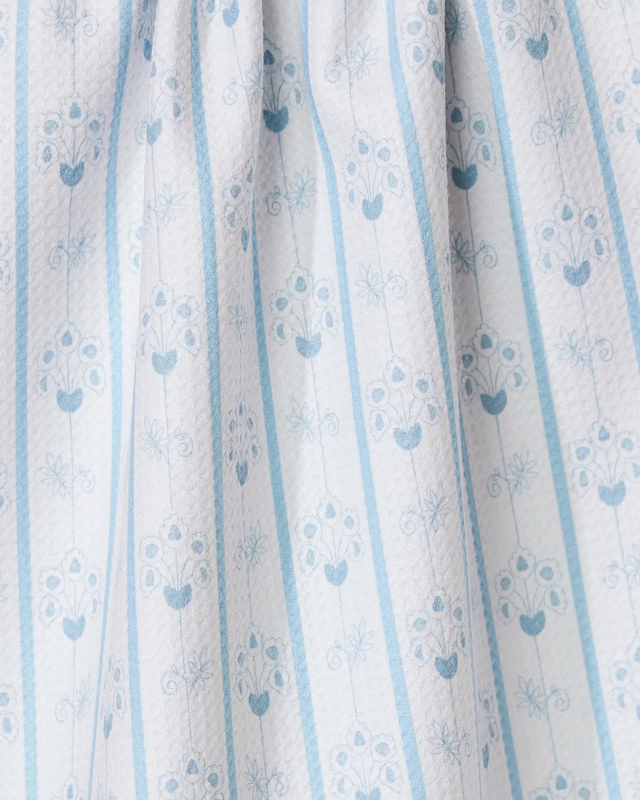 A close-up of GEOMETRY’s Ediths English Cottage recycled kitchen towel shows white, pleated fabric with a textured surface, vertical blue stripes, and a repeating light blue floral and leaf pattern.