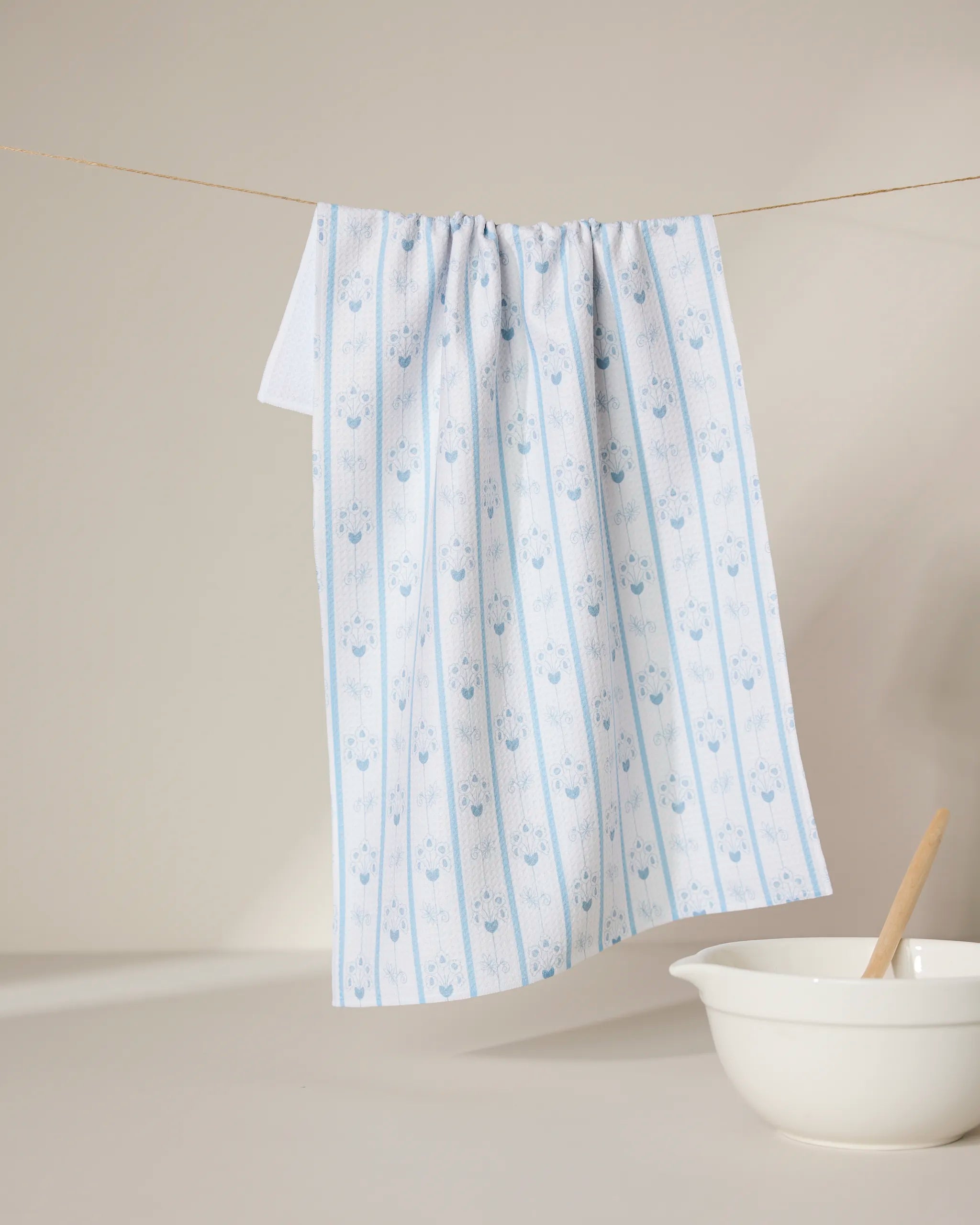 Ediths English Cottage tea towel by GEOMETRY, featuring light blue florals and stripes, hangs beside a white bowl and wooden spoon—its quick-drying fabric makes it ideal for busy kitchens.