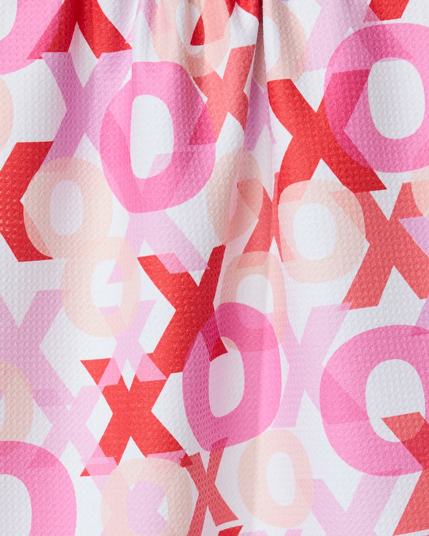A close-up of the GEOMETRY Embrace towel, featuring ultra-absorbent, textured fabric with overlapping X and O patterns in red, pink, and light peach on a white background.
