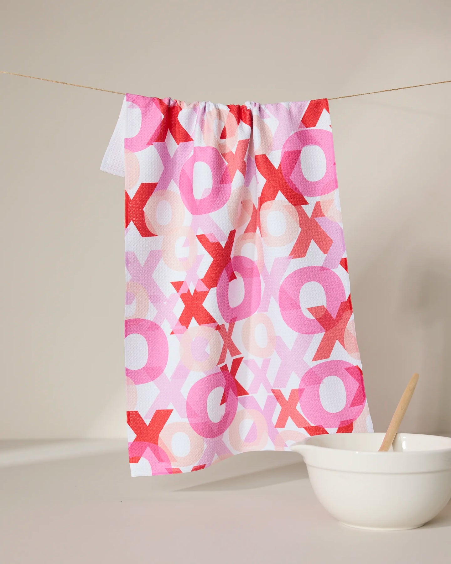 The GEOMETRY Embrace recycled white kitchen towel, featuring pink, red, and peach XOXO patterns, is displayed hanging on a string above a white bowl with a wooden spoon against a neutral background.