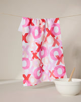 The GEOMETRY Embrace recycled white kitchen towel, featuring pink, red, and peach XOXO patterns, is displayed hanging on a string above a white bowl with a wooden spoon against a neutral background.