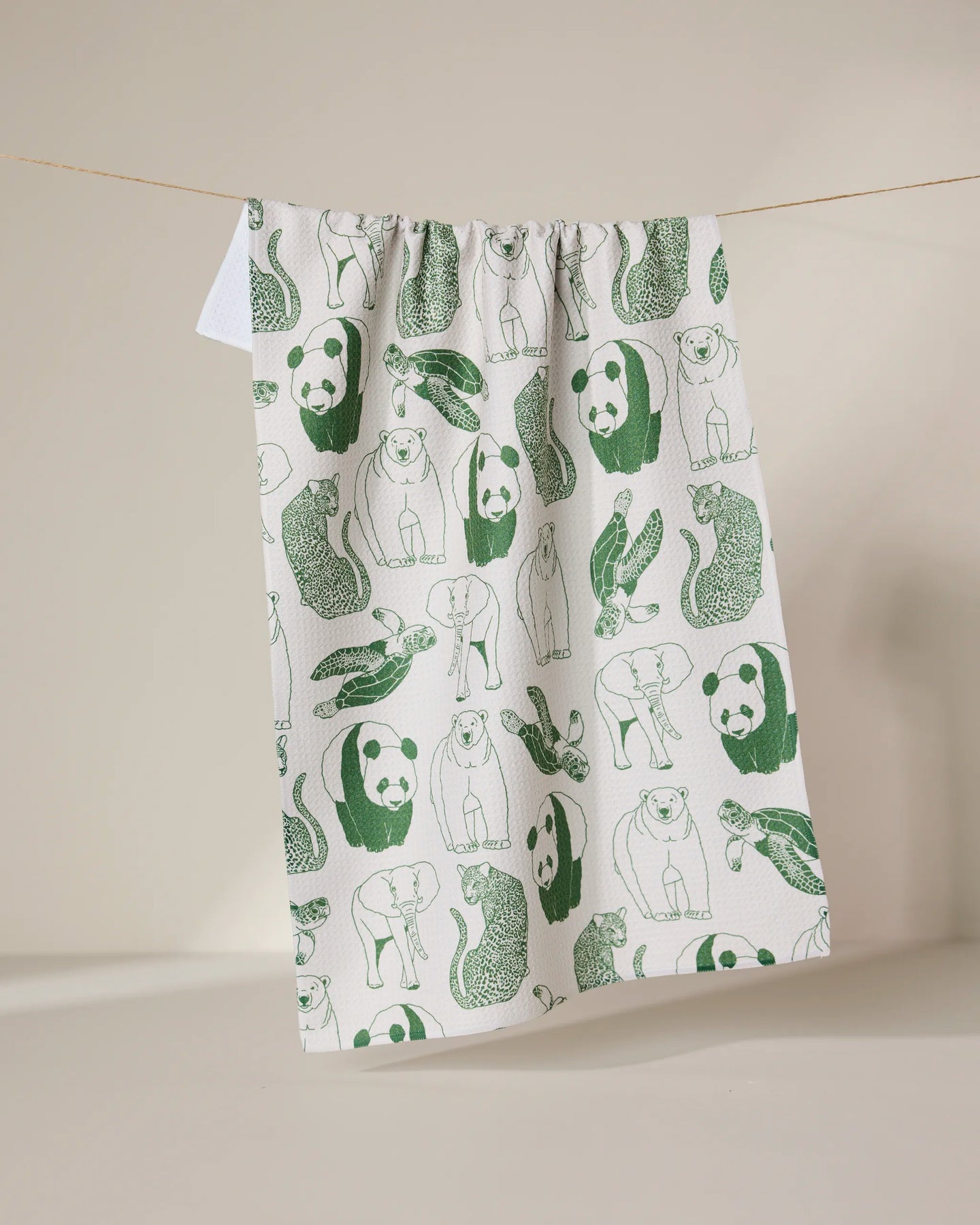 The GEOMETRY Endangered tea towel, made from recycled materials, dries quickly and features green illustrations of pandas, snakes, rabbits, and otters. It hangs on a clothesline against a neutral background.