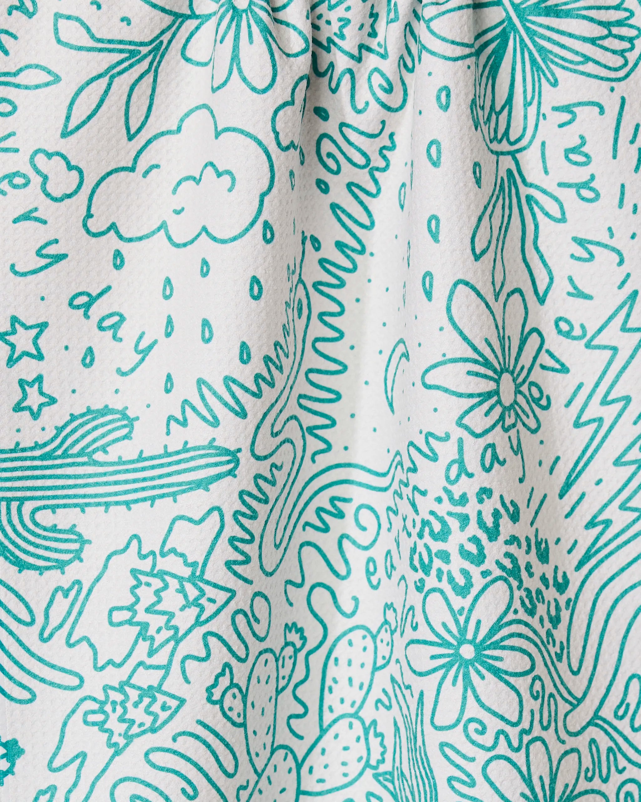 The GEOMETRY Every Day tea towel features a white textured design with teal doodles—flowers, clouds, stars, rain, cacti, and squiggles—plus the phrase every day repeated across its ultra-absorbent surface.