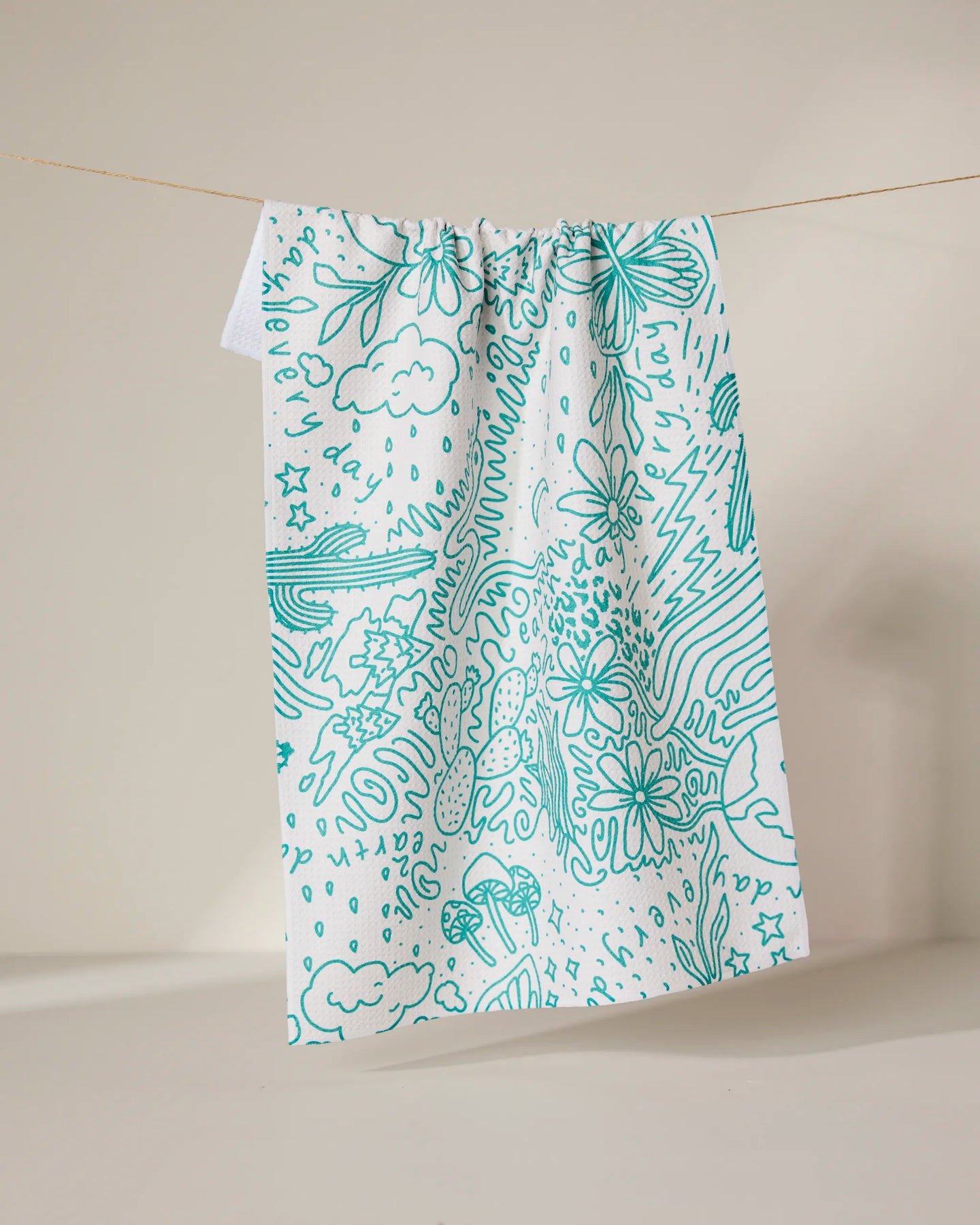 The Every Day tea towel by GEOMETRY, made from recycled materials, displays teal line art of flowers, mushrooms, clouds, stars, and abstract shapes on white fabric, shown hanging against a neutral backdrop.