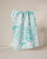 The Every Day tea towel by GEOMETRY, made from recycled materials, displays teal line art of flowers, mushrooms, clouds, stars, and abstract shapes on white fabric, shown hanging against a neutral backdrop.