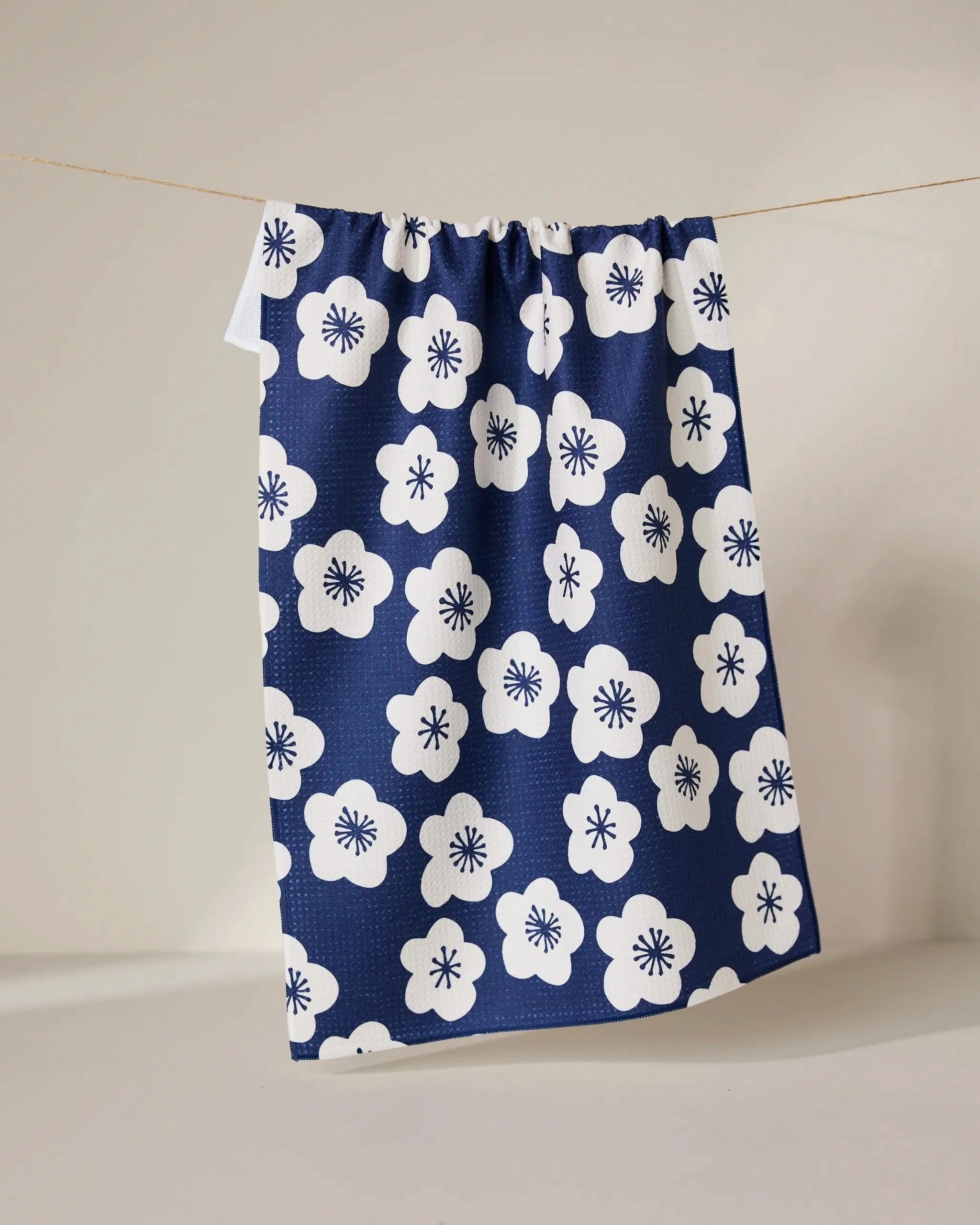 The GEOMETRY Every Sunday blue eco-friendly tea towel featuring a white floral pattern hangs on a string against a neutral background.