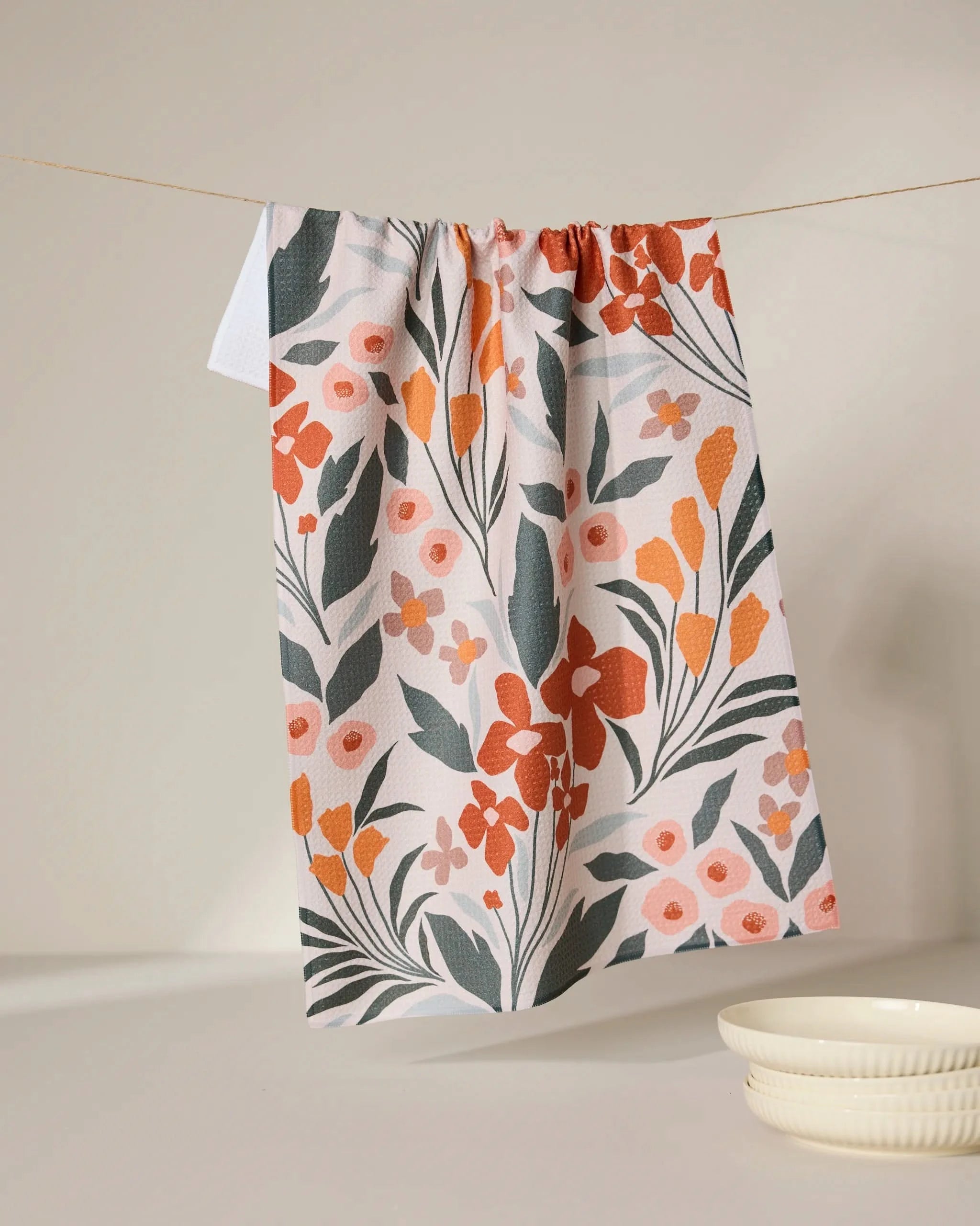 The Fallowers tea towel by GEOMETRY, crafted from recycled materials and featuring orange, pink, and green botanical patterns, hangs above a white surface beside two stacked white dishes.
