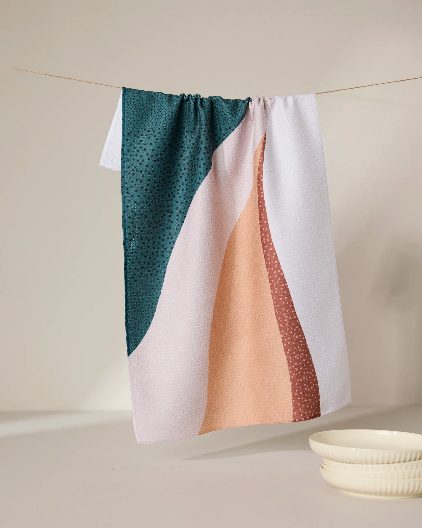 The GEOMETRY Fan kitchen towel features abstract wavy patterns in dark green, peach, and rust. Made from recycled materials for eco-friendly style, it hangs above a white ceramic dish and bowl on a neutral background.